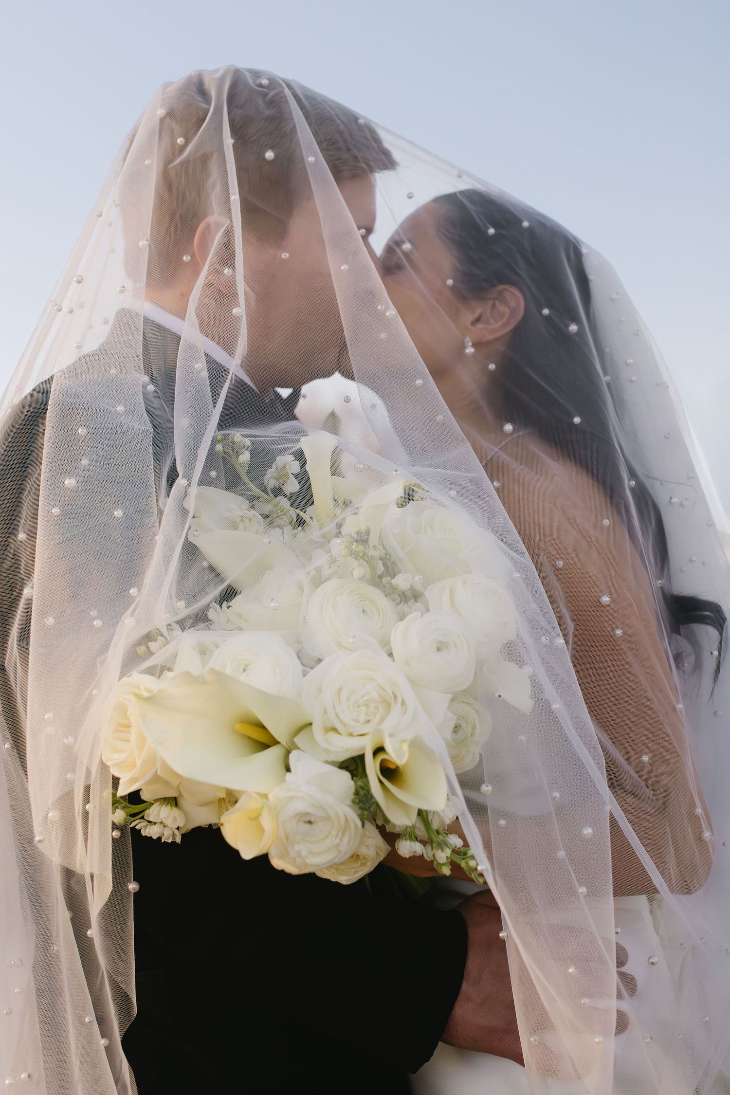 Romantic close-up of couple under wedding veil with white rose bouquet.