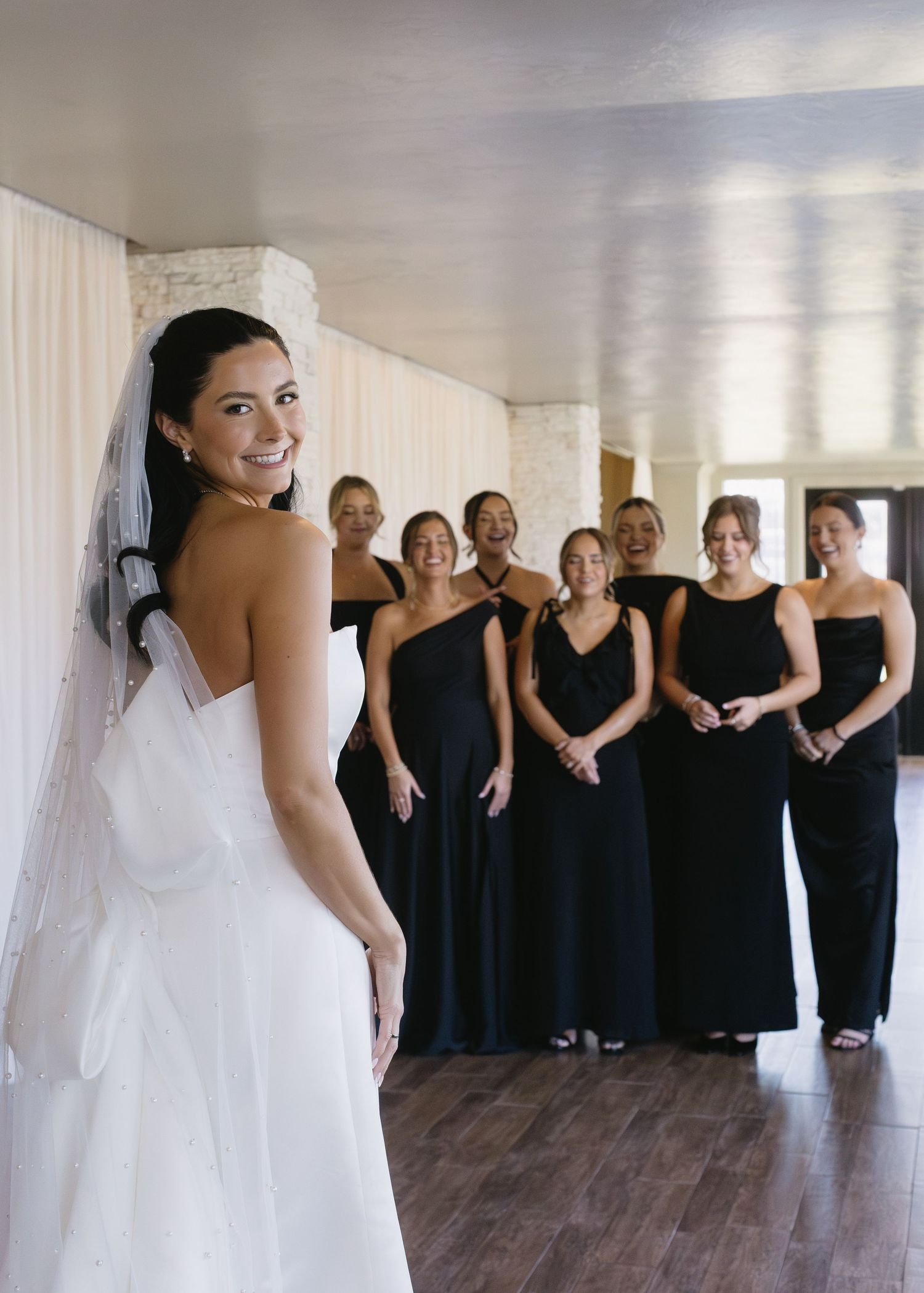 Bridesmaids in black dresses react with joy upon seeing the bride in her wedding gown.