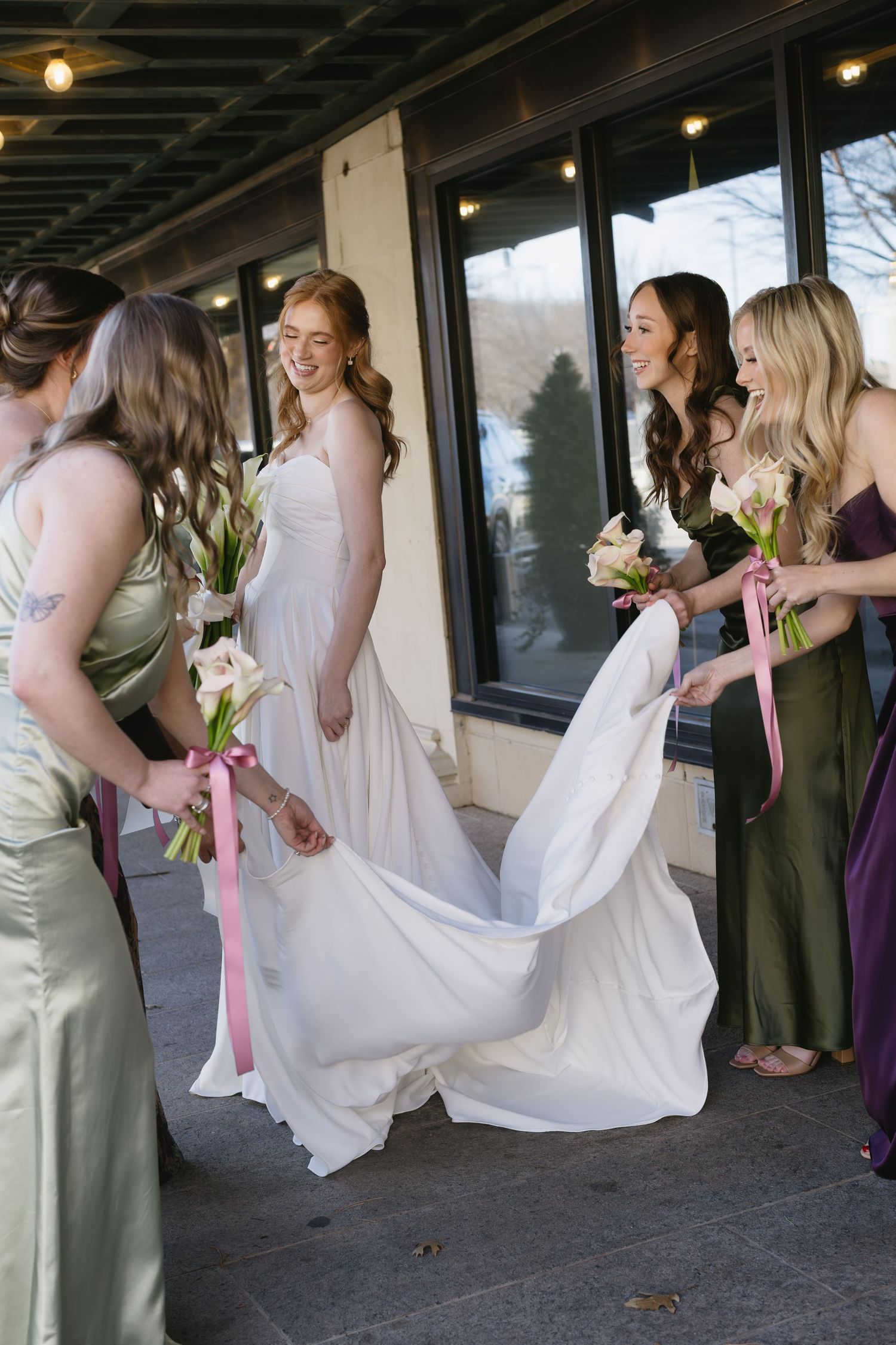 Bridesmaids in various colored dresses playfully lifting their skirts in an outdoor venue.
