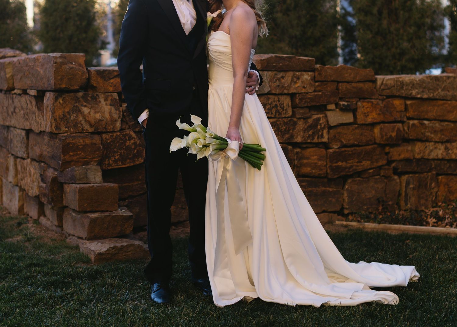Elegant wedding couple poses by rustic stone wall during golden hour.