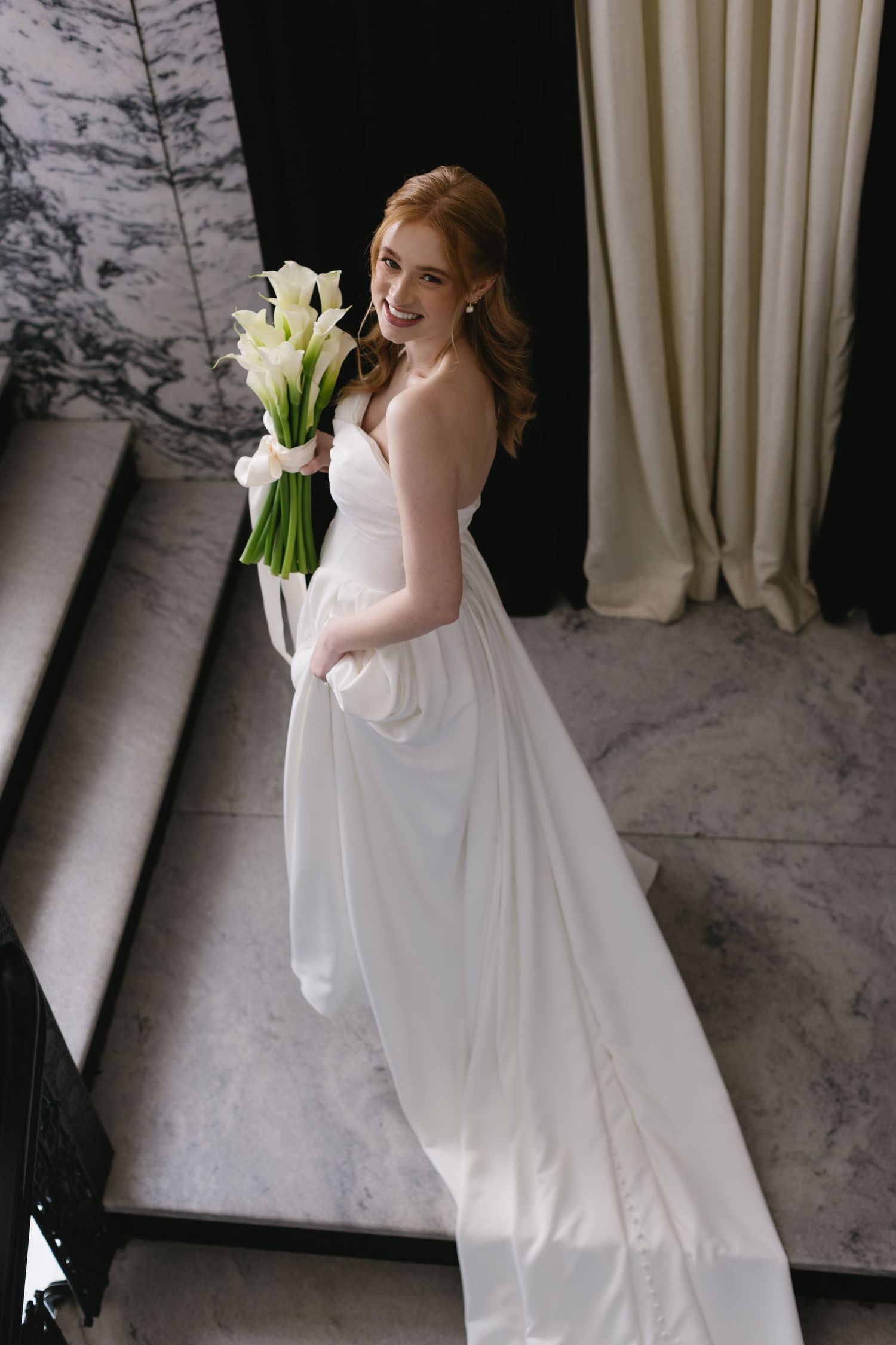 Elegant bride in white gown holding a bouquet of calla lilies against marble wall.
