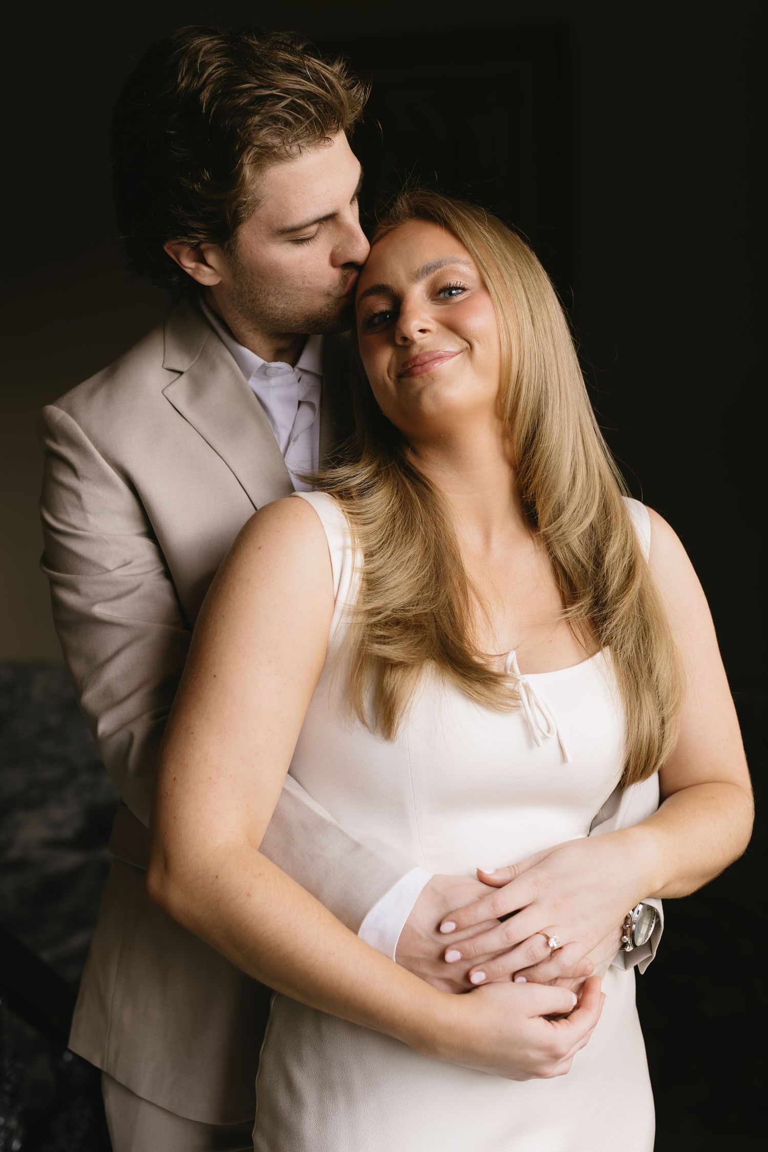 A romantic couple embraces in a tender moment, with the woman wearing white and the man in a beige suit.