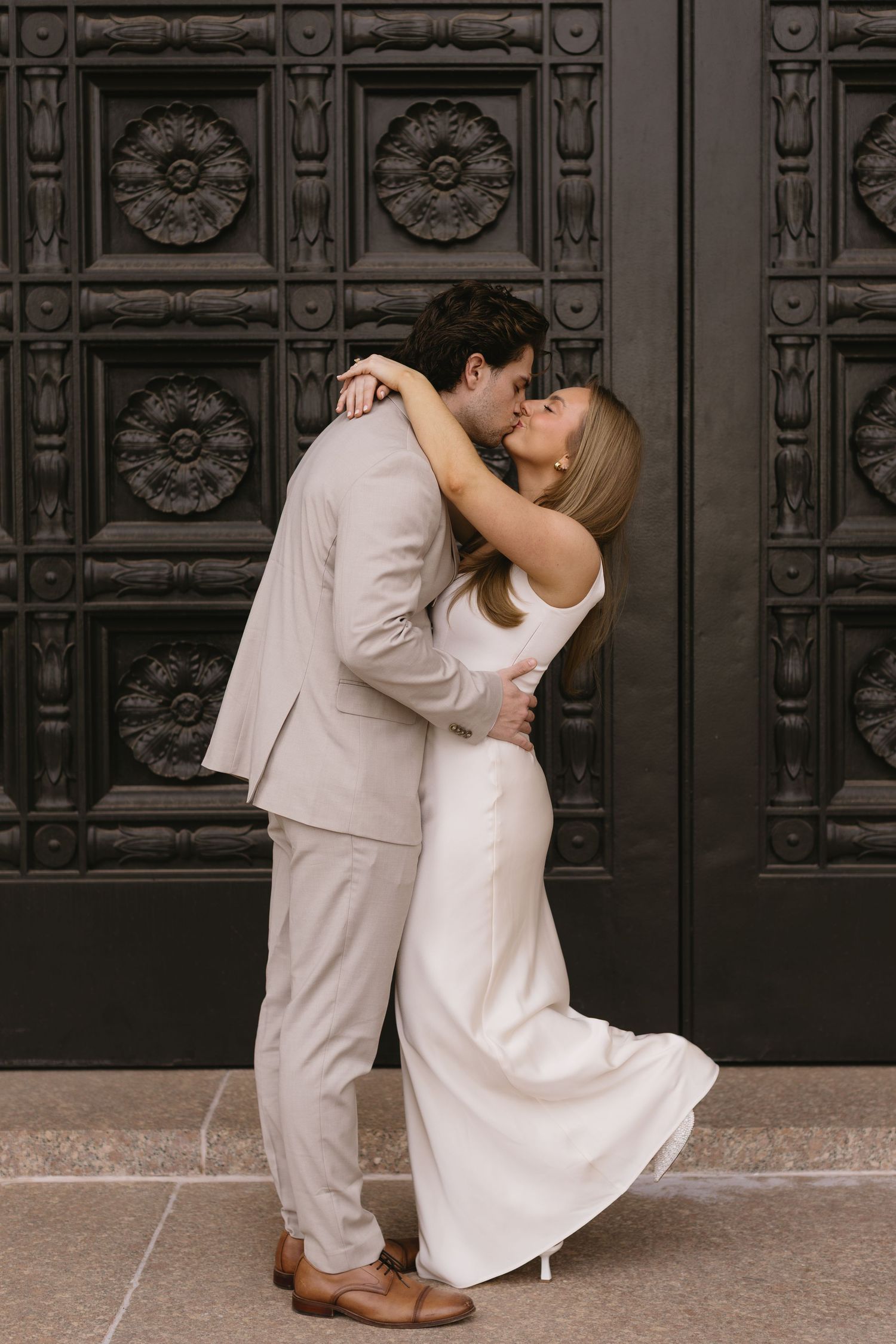 A couple shares a romantic kiss in front of an ornate black door with decorative floral patterns.