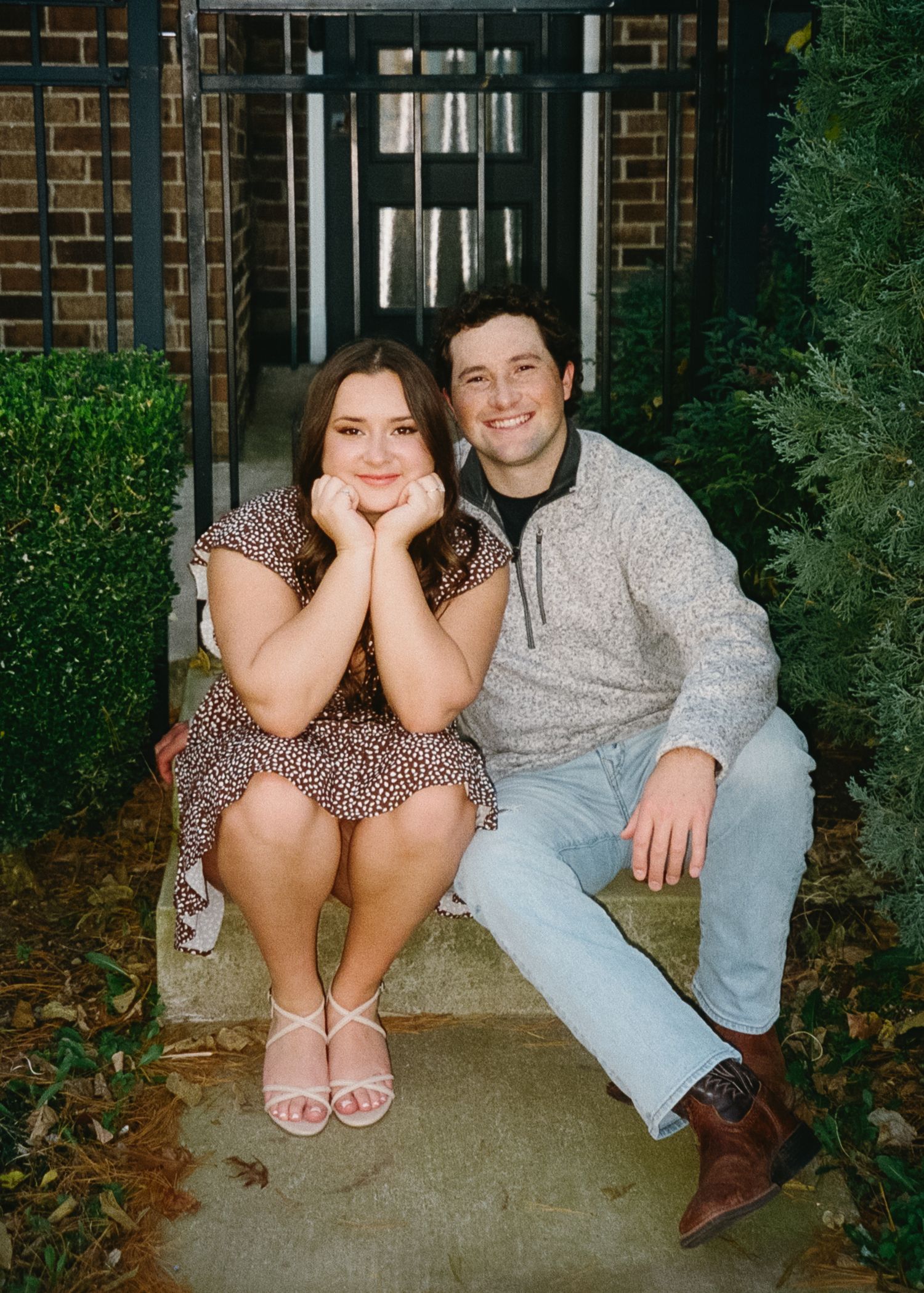 A couple sits together on a concrete porch step in a casual outdoor setting with brick walls and greenery.