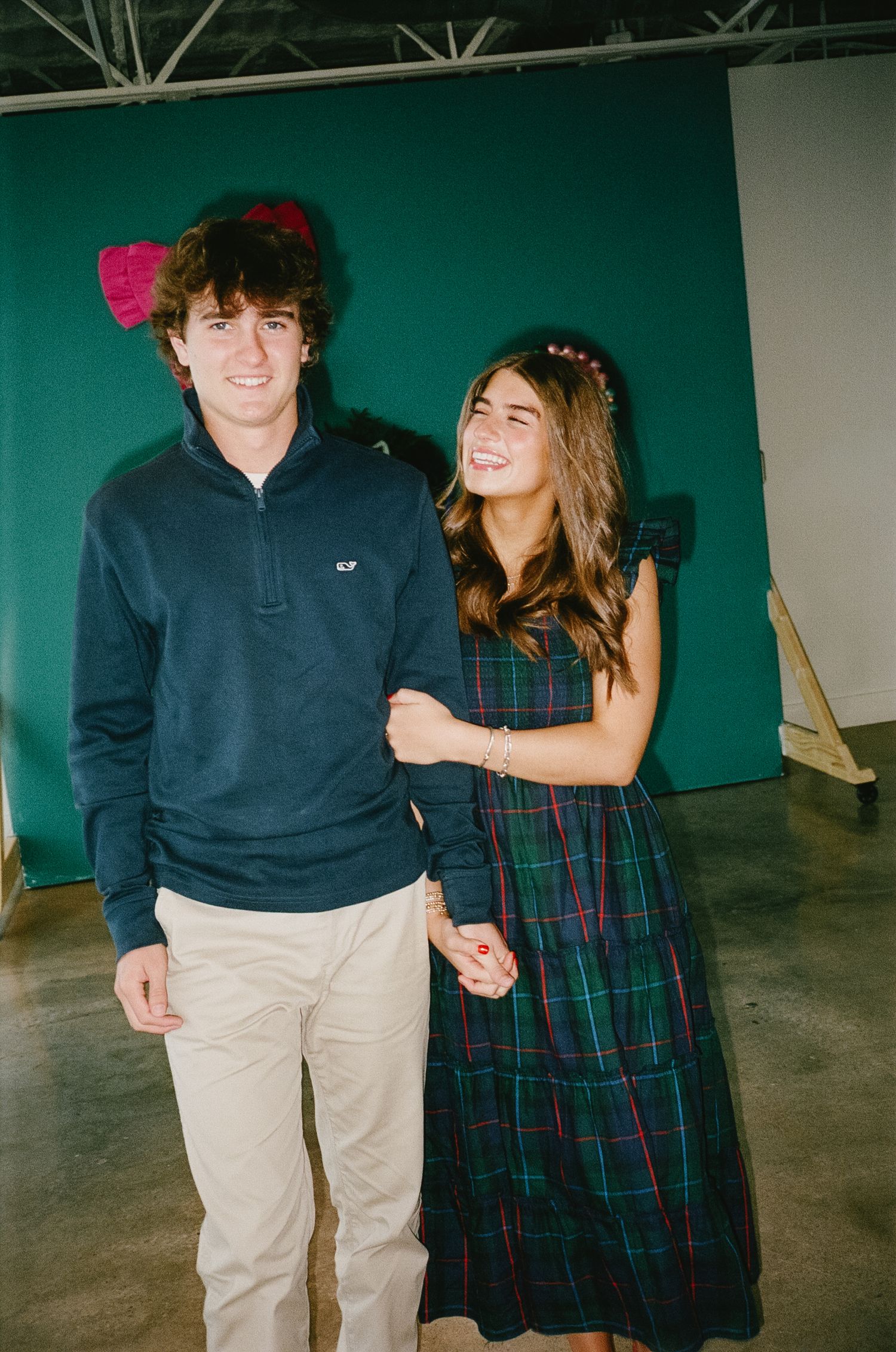 A couple poses together against a teal backdrop, wearing coordinated navy and plaid holiday outfits.