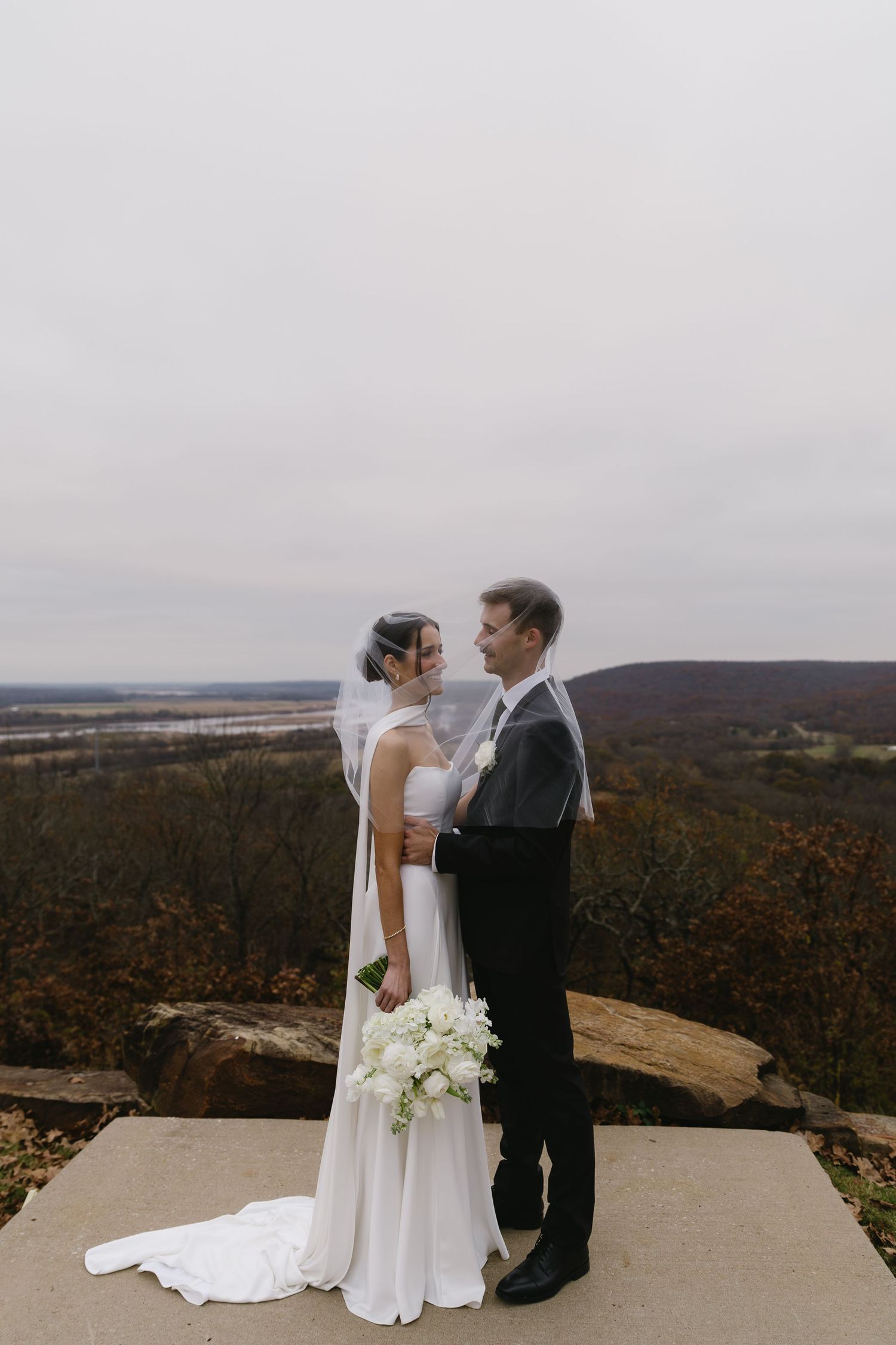 Couple in wedding attire poses on scenic overlook with autumn landscape in background.