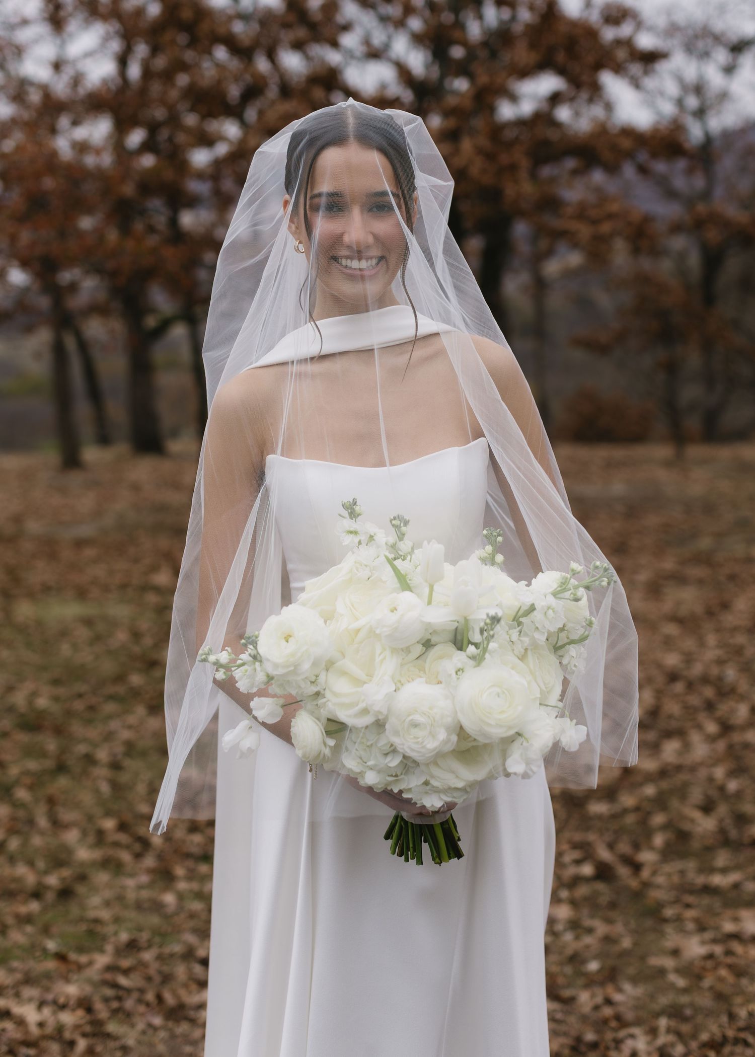 A bride in a white wedding dress and veil holds a bouquet of white roses against an autumn backdrop.