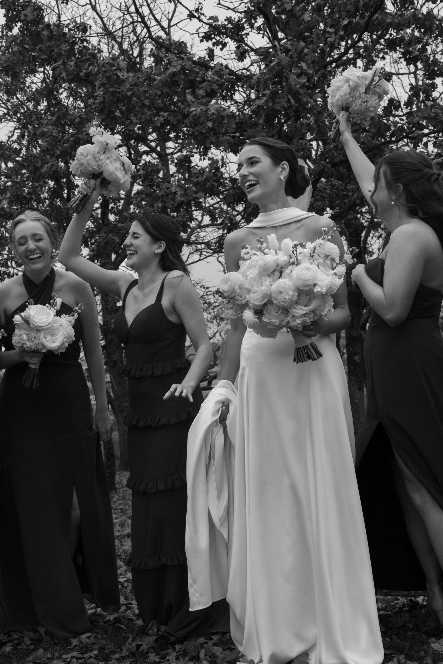 Wedding party laughs and celebrates together outdoors with bridesmaids in black and bride in white.