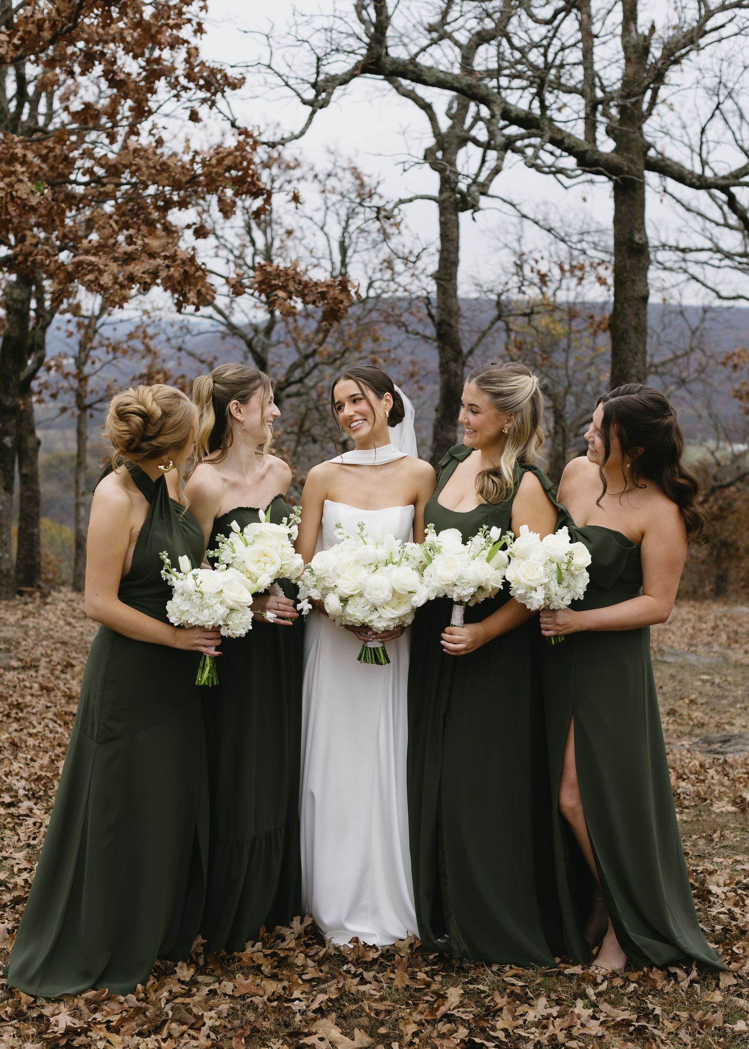 A bridal party in dark green bridesmaid dresses and white bouquets poses together on fallen autumn leaves.
