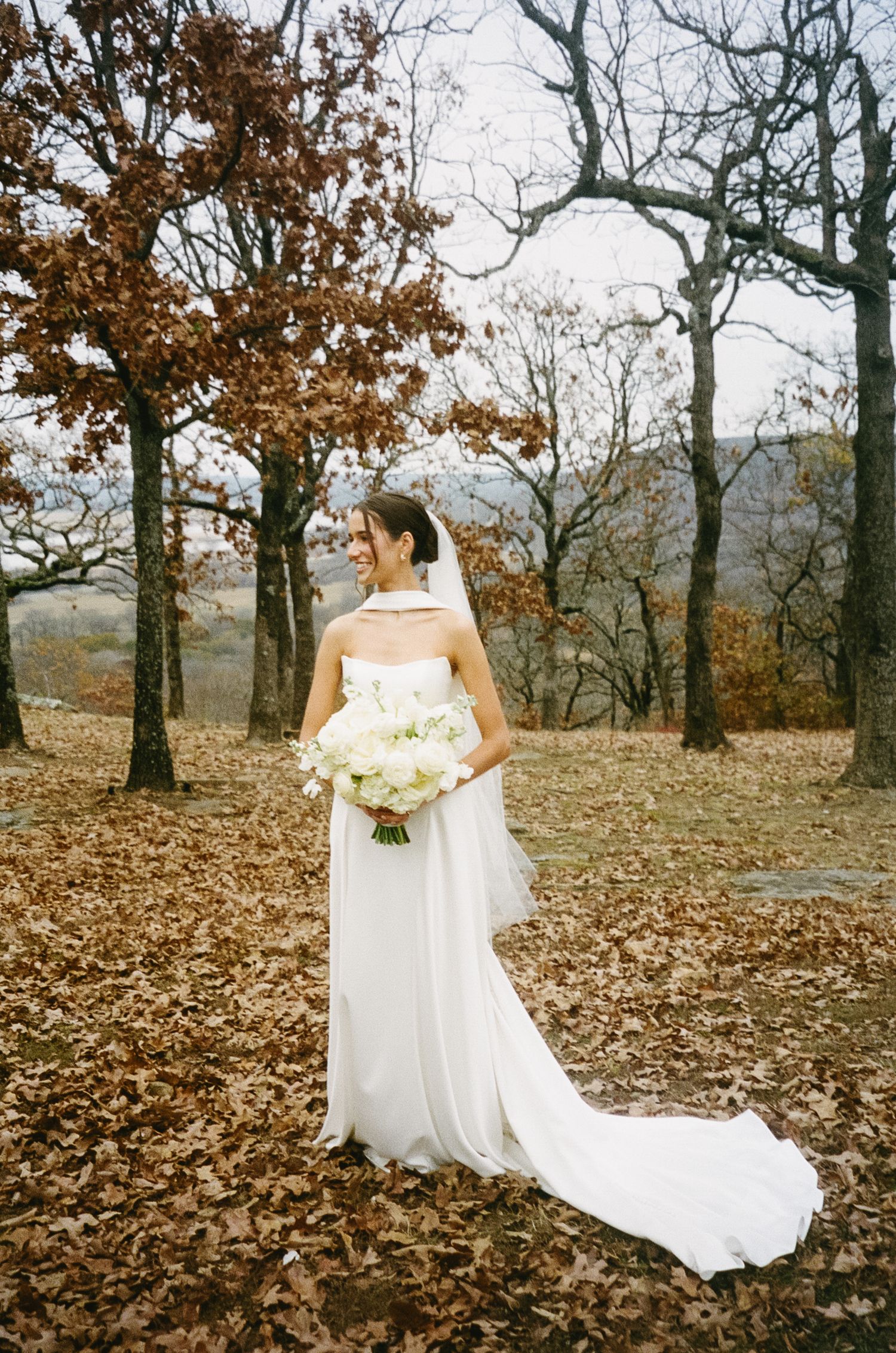 Bride holding white bouquet among autumn trees with fallen leaves.