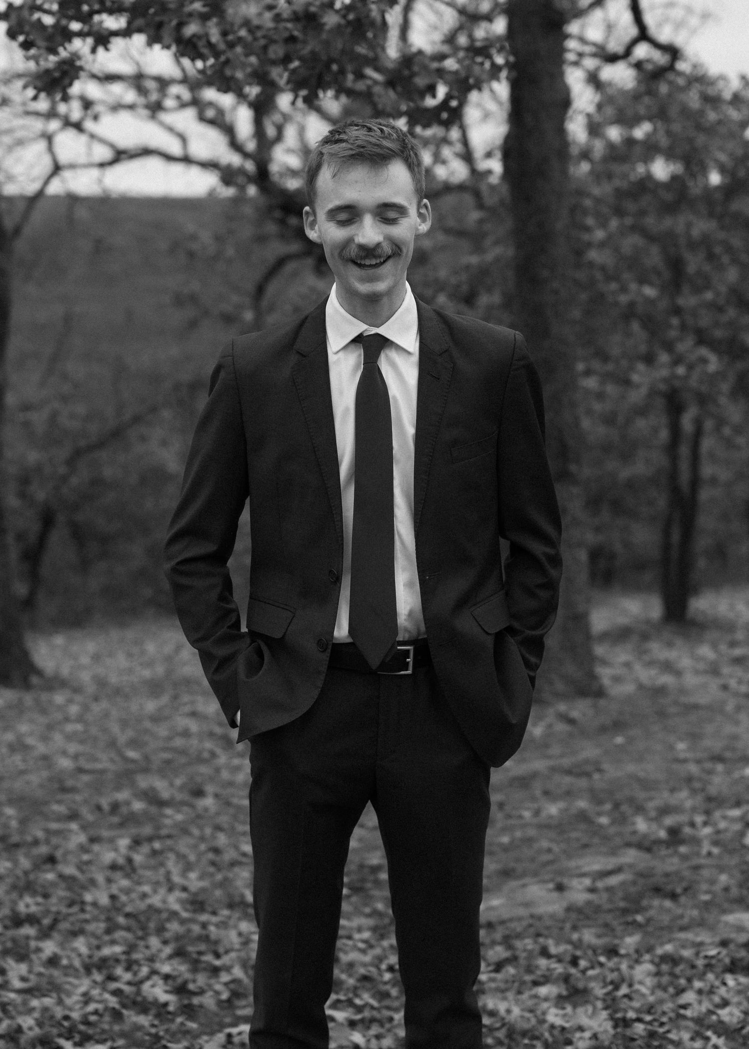 Black and white portrait of someone in a suit standing outdoors among autumn trees.