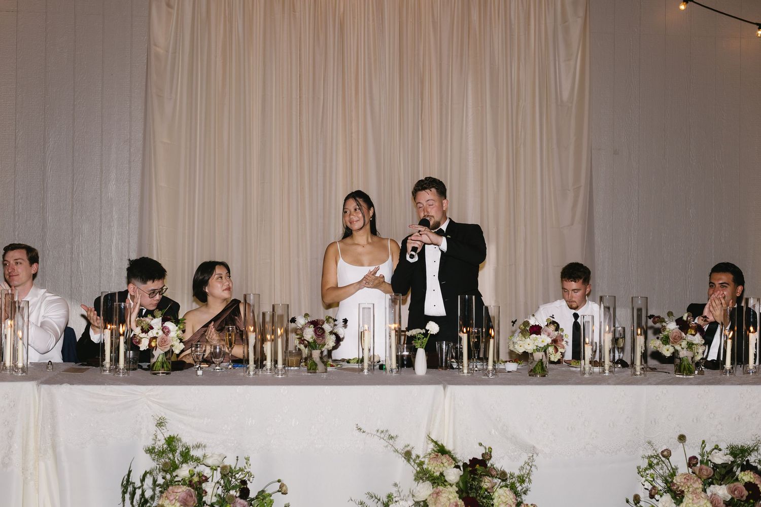 Wedding reception hall decorated with white drapes and floral centerpieces on dining tables.