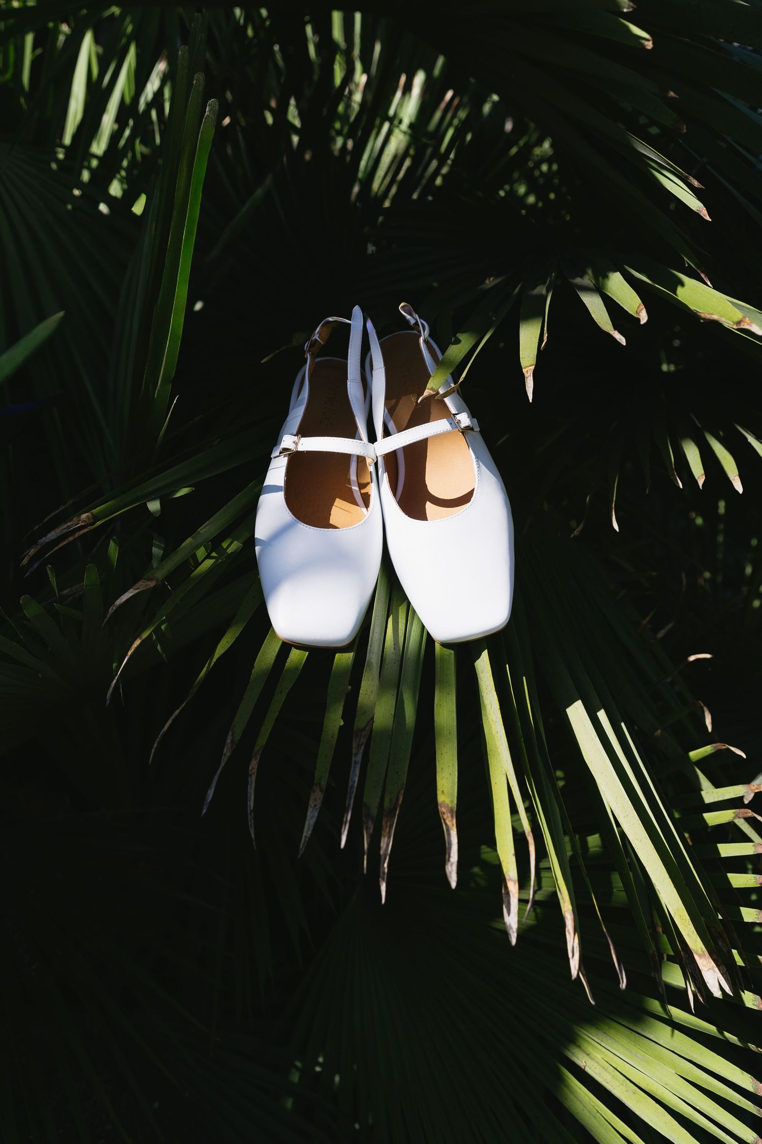 White wedding shoes displayed against tropical palm fronds in natural lighting.
