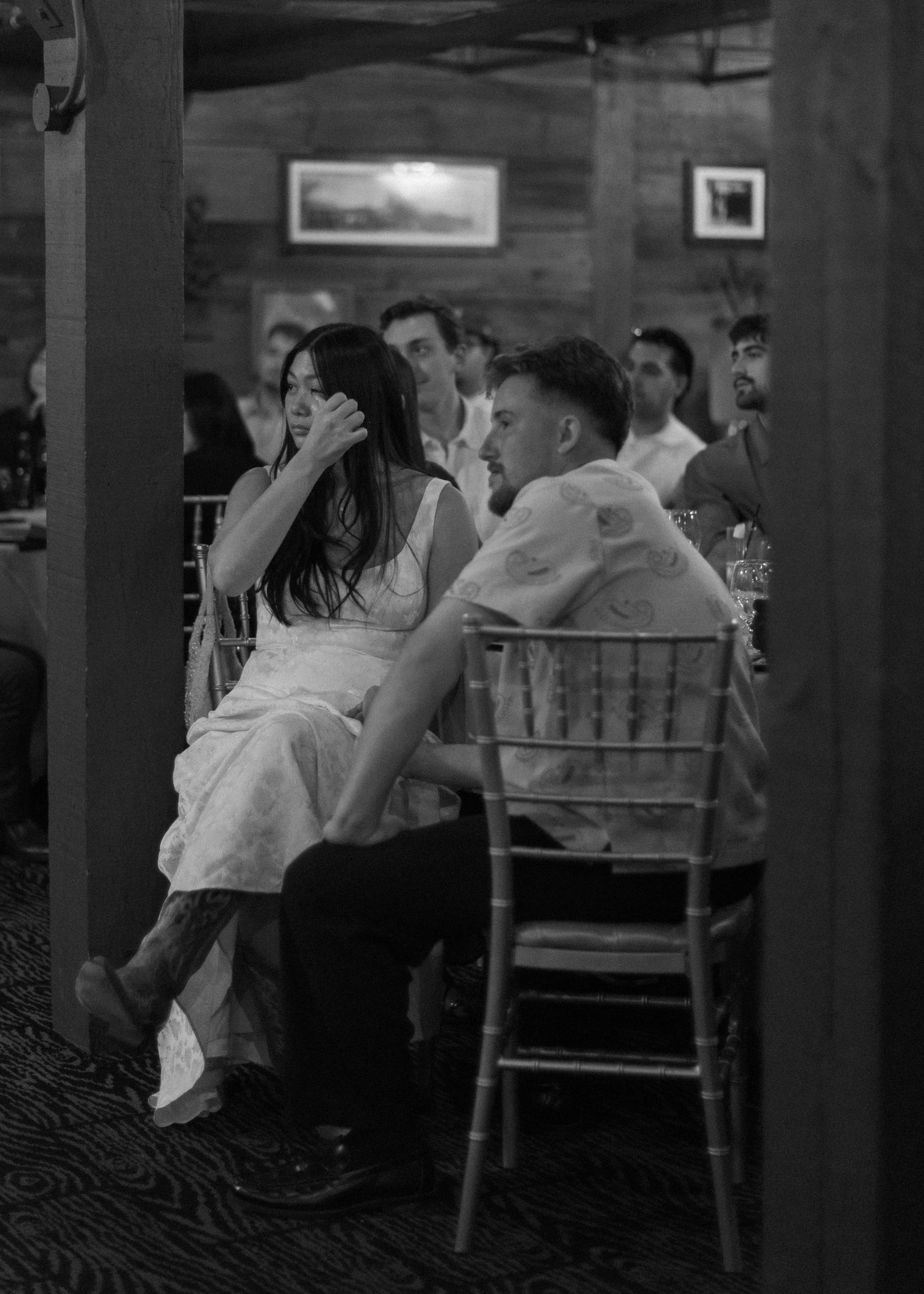 Wedding reception guests seated at tables in an indoor venue with wooden chairs and dim lighting.