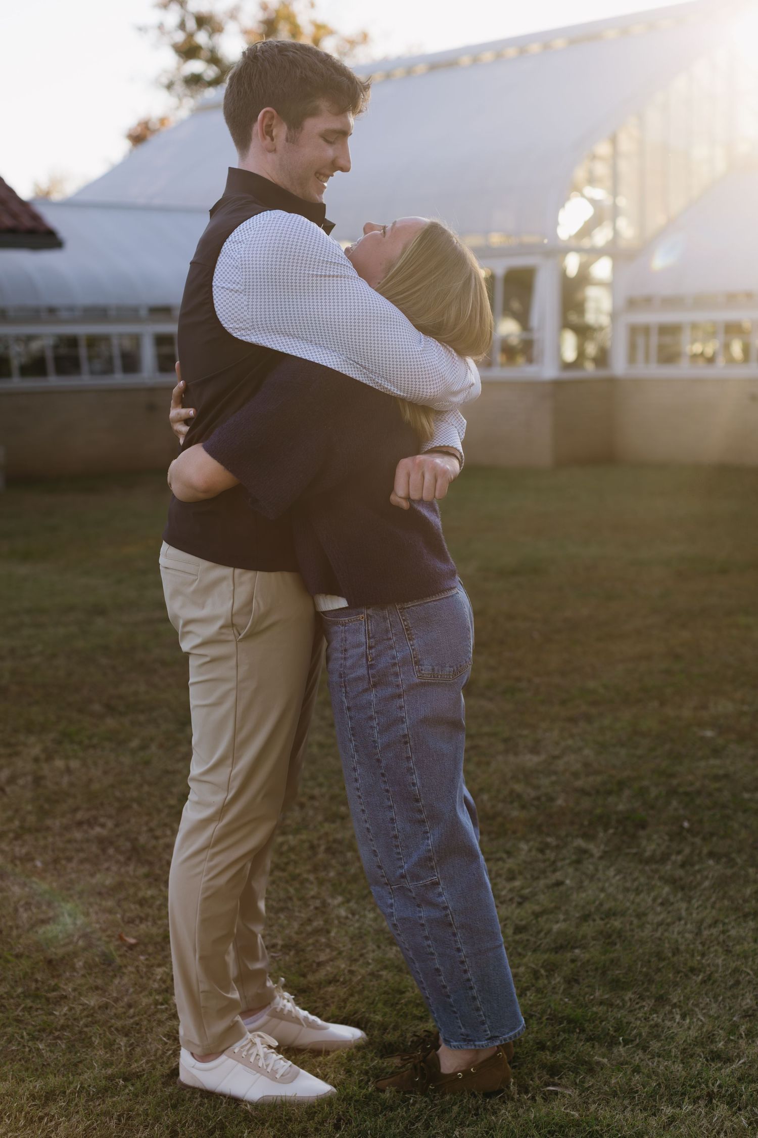 A couple embraces in a grassy field at sunset wearing casual clothing and white sneakers.