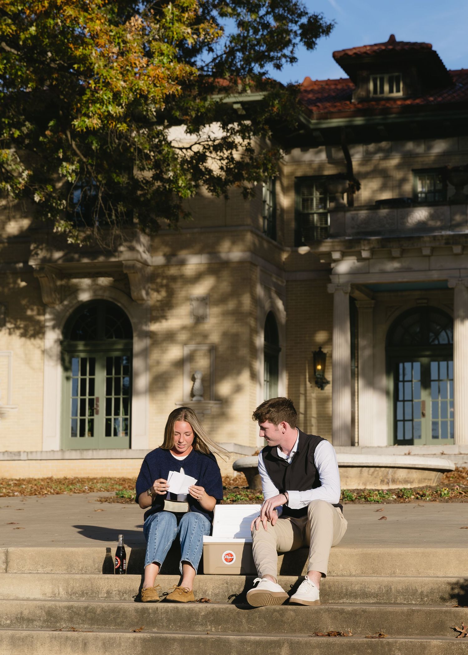 A couple sits casually on stone steps outside a building with autumn trees in background.