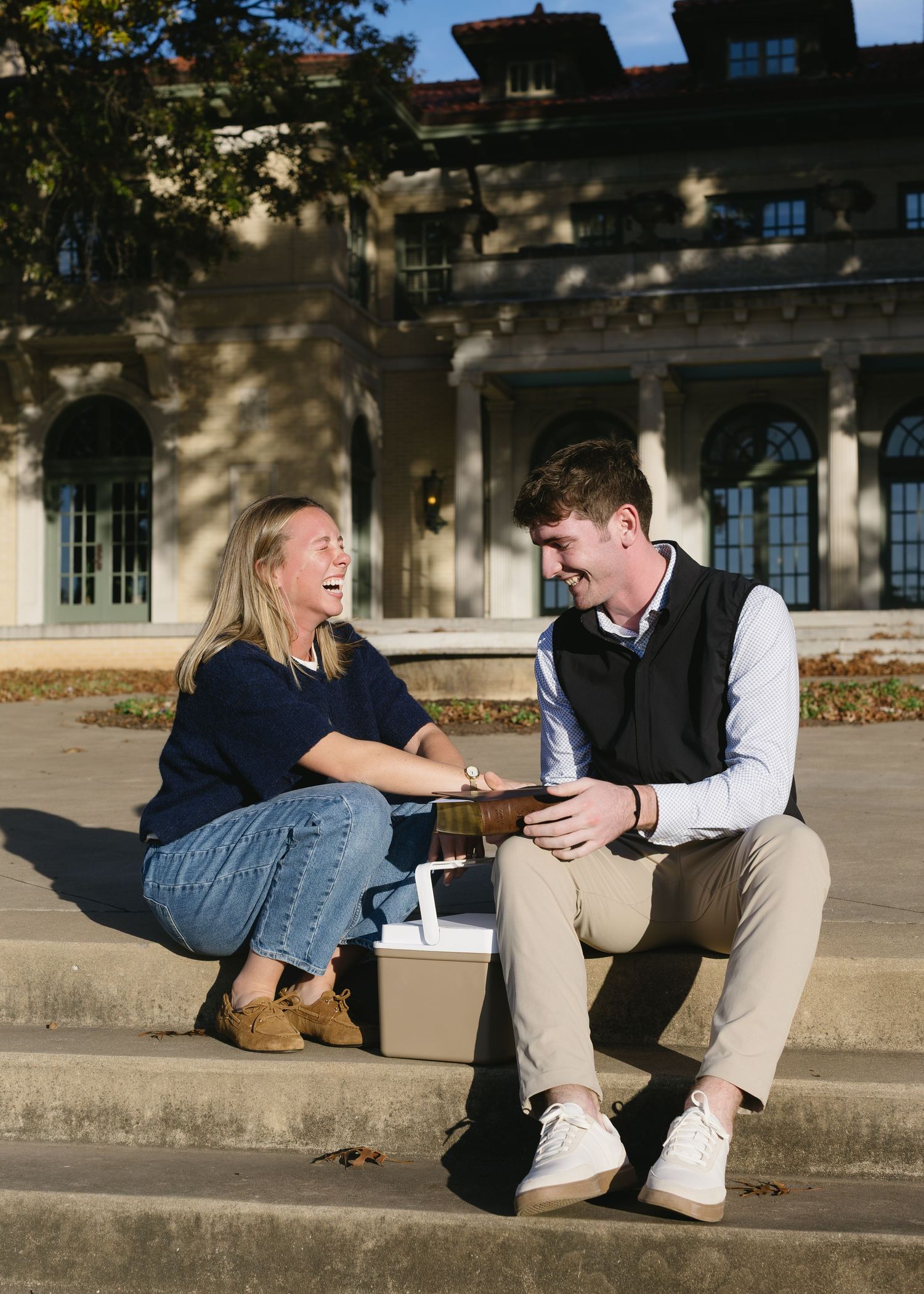 Two people sitting and talking on concrete steps in front of a classic building.