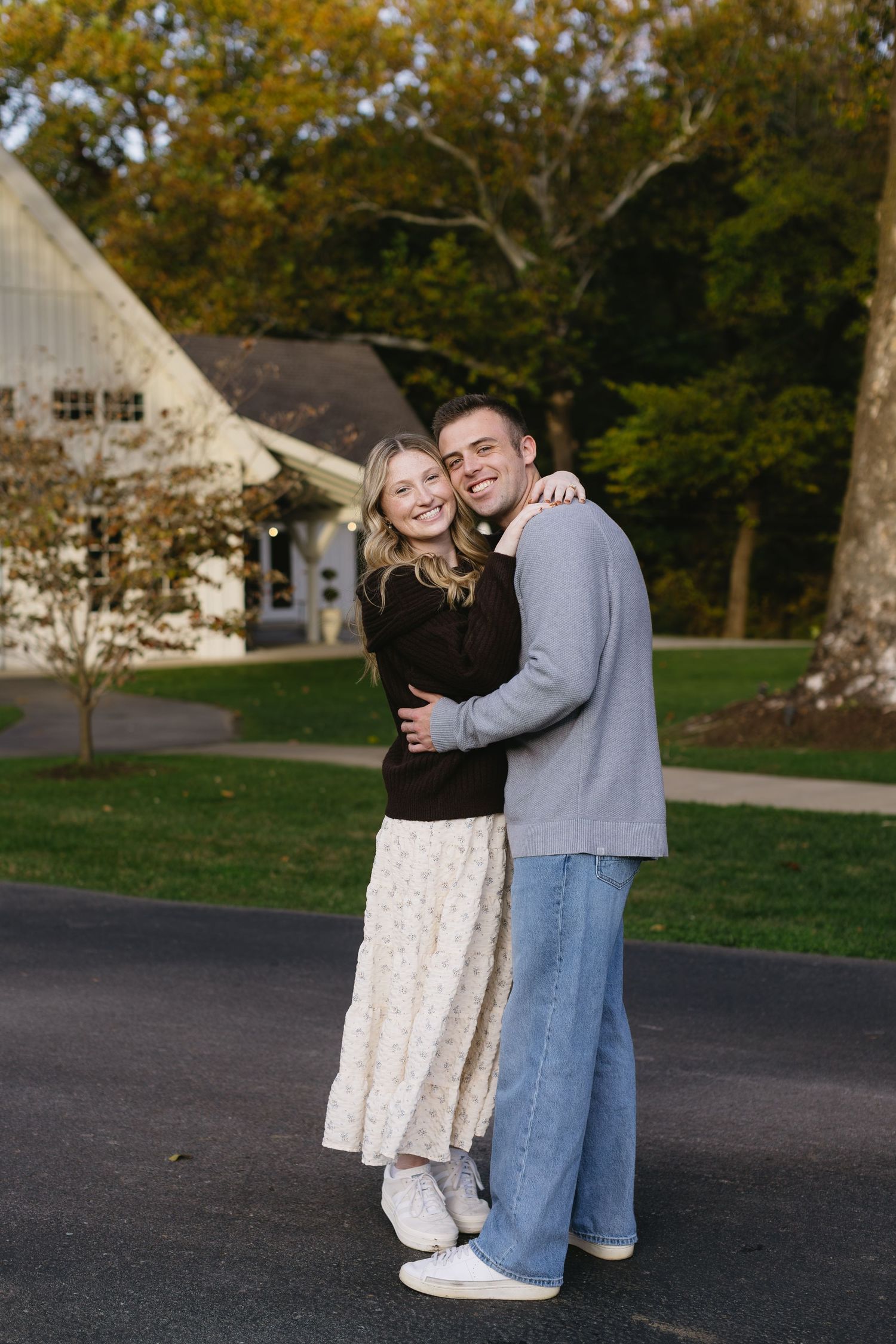 A couple embraces and smiles together outside a white barn building in autumn.