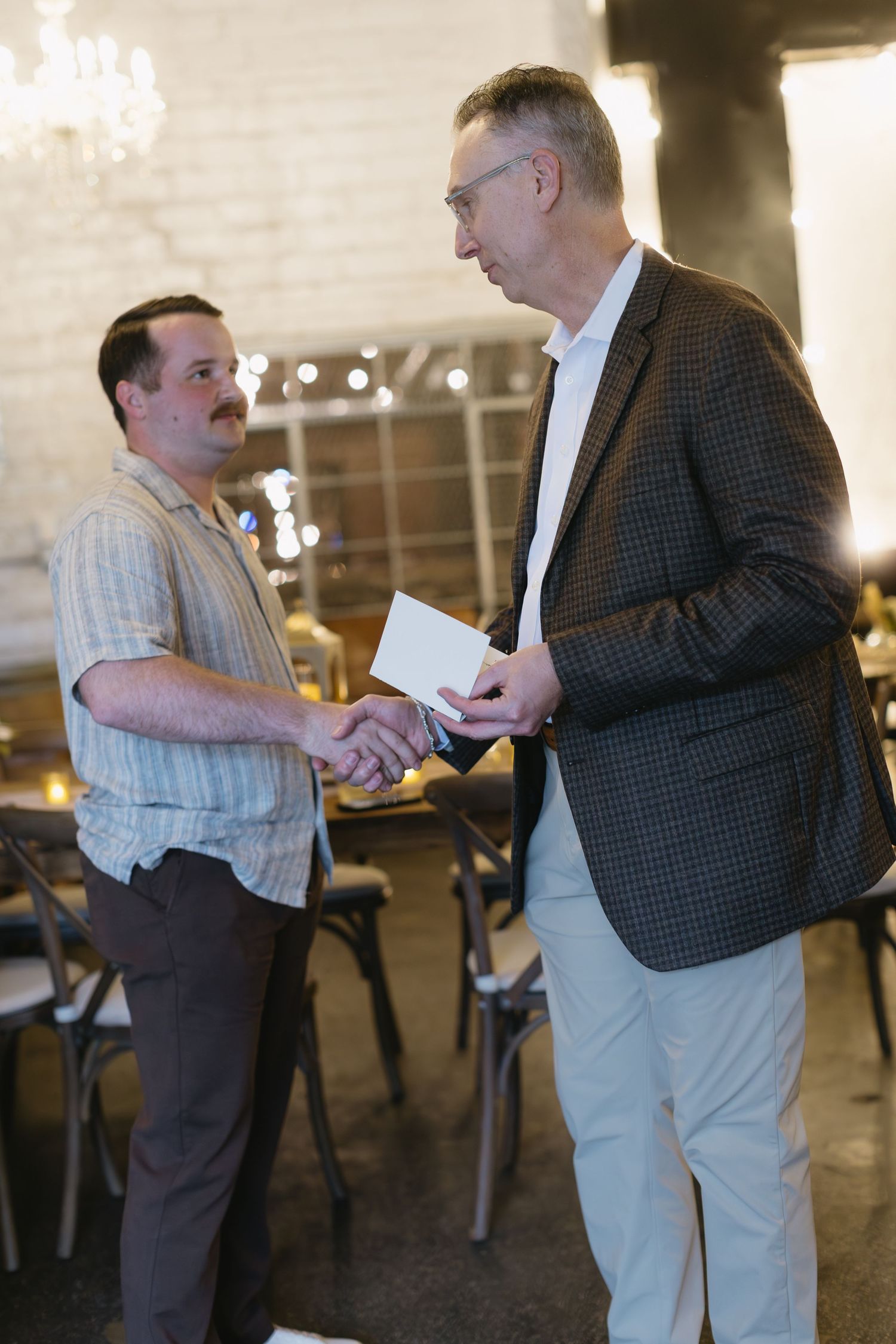 Two people shaking hands and exchanging documents in an event space with chandeliers.
