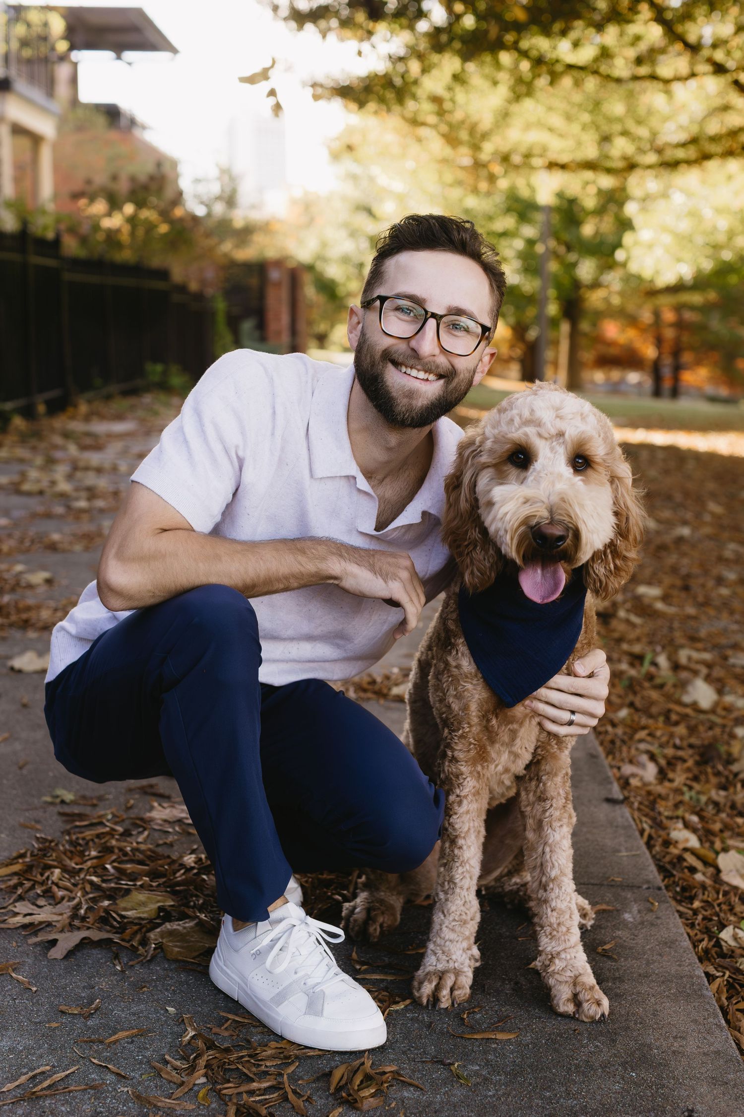 A smiling person in white shirt and blue pants poses with their goldendoodle dog on fallen autumn leaves.