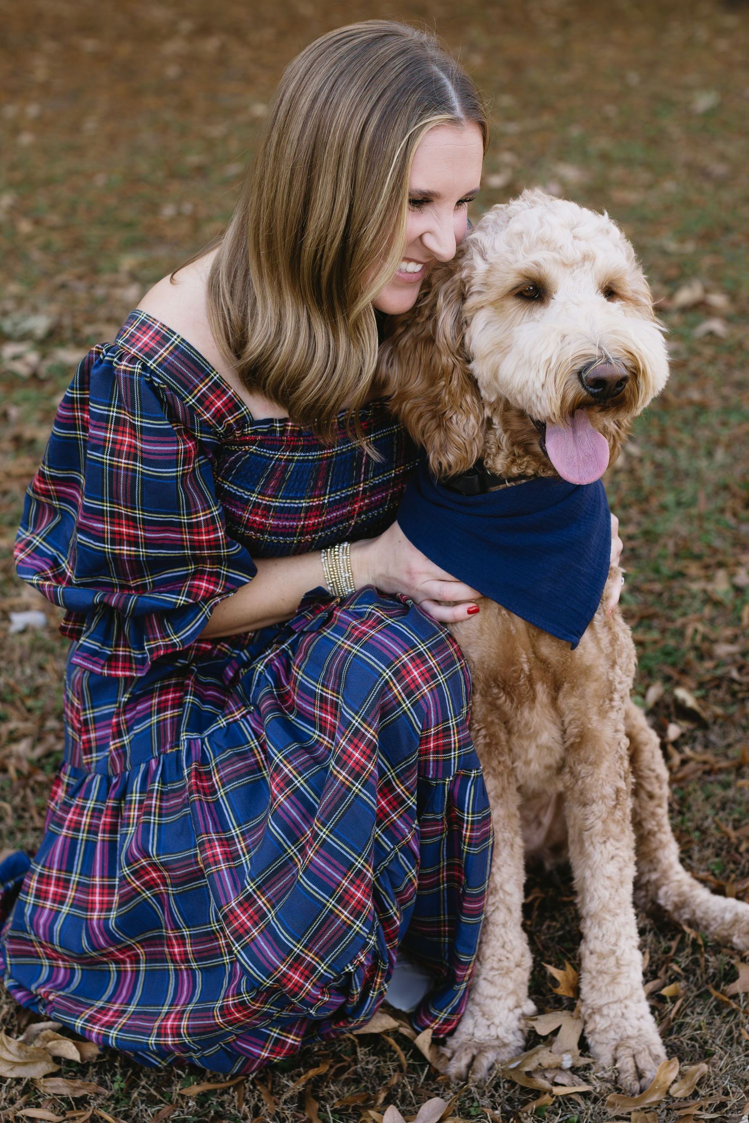 A fluffy goldendoodle dog sits with someone wearing a plaid dress in a park during fall.