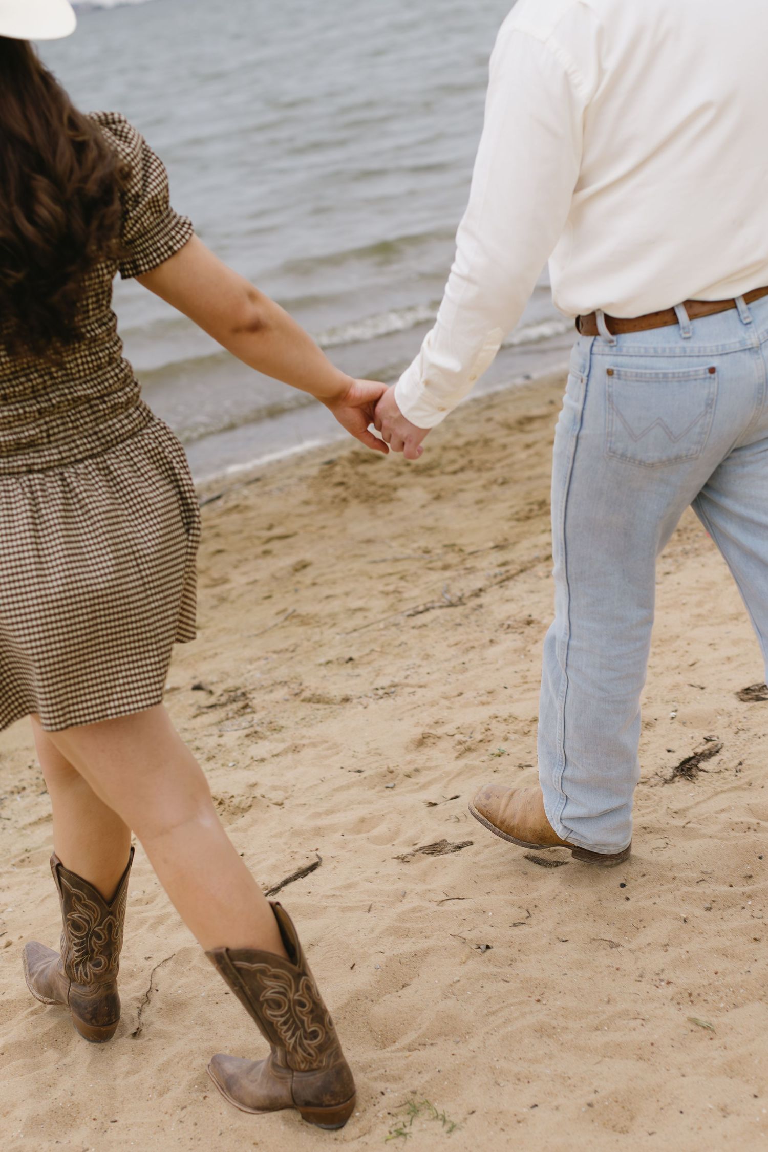 Couple walking hand in hand on the beach, focusing on their boots and footprints in the sand.