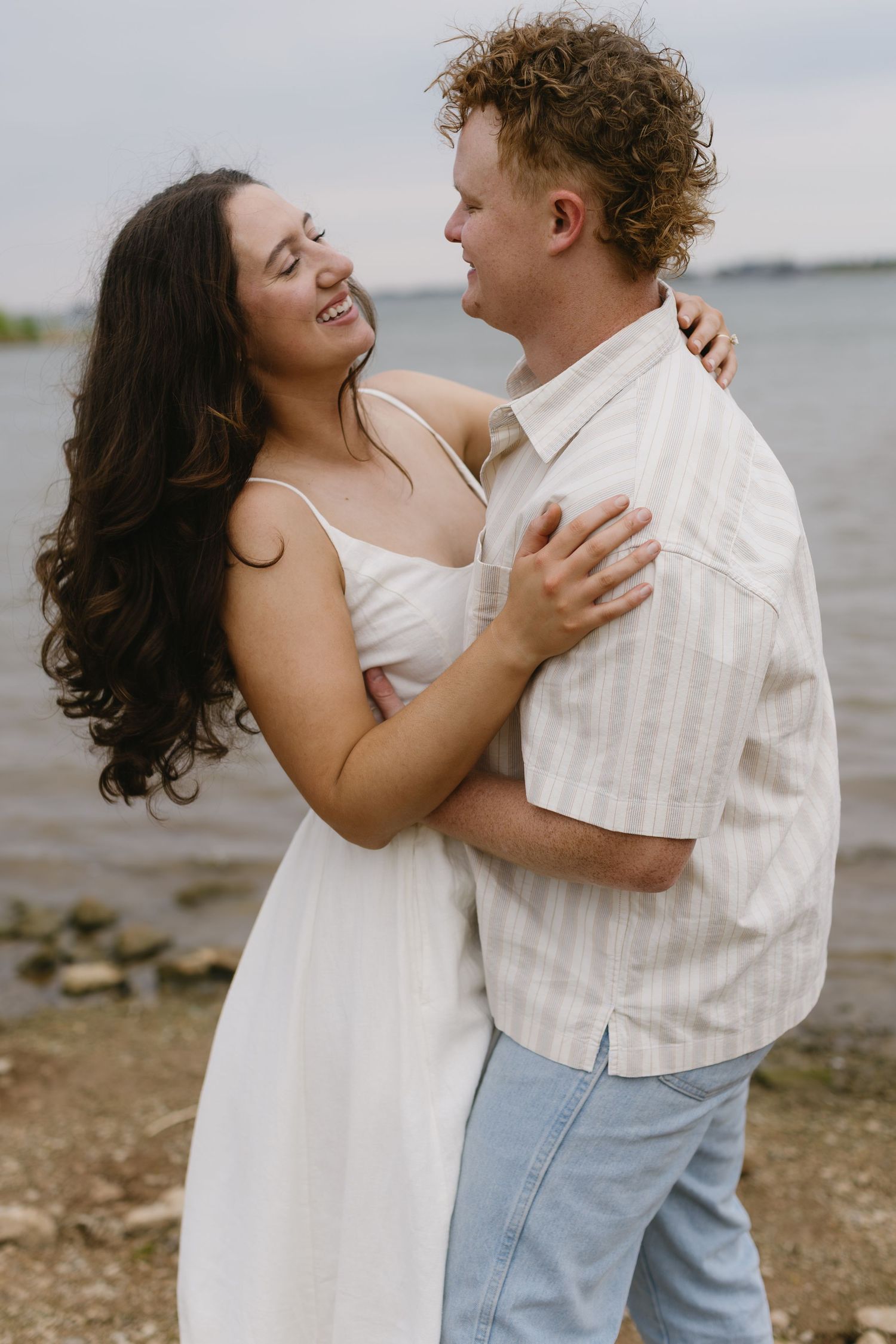 Romantic couple embraces by the waterfront, dressed in white and beige, sharing intimate moments.
