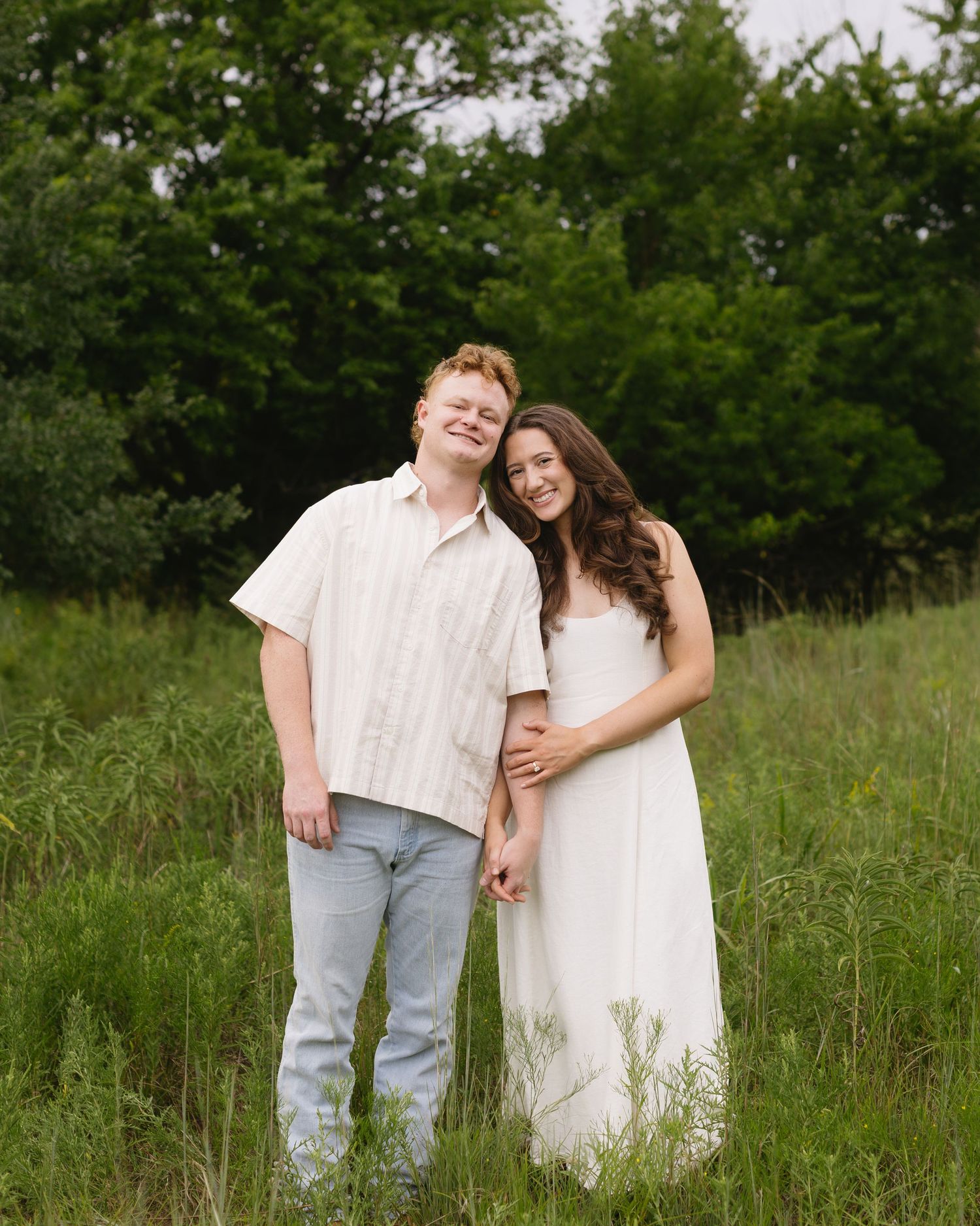 A couple stands affectionately in a sun-lit meadow with tall grass and trees in the background.