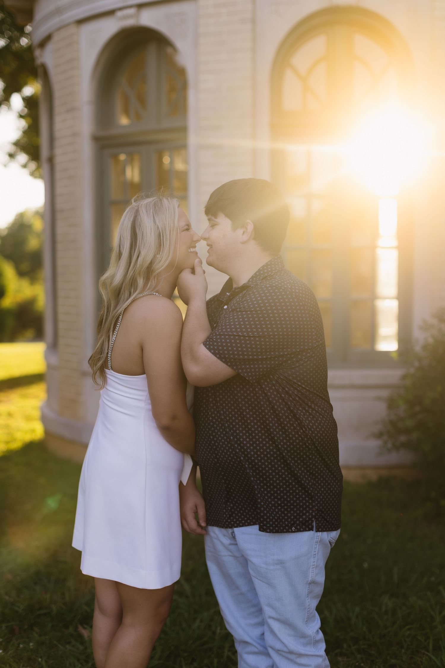 Romantic couple sharing an intimate moment outside a building at golden hour with sun flare in the background.