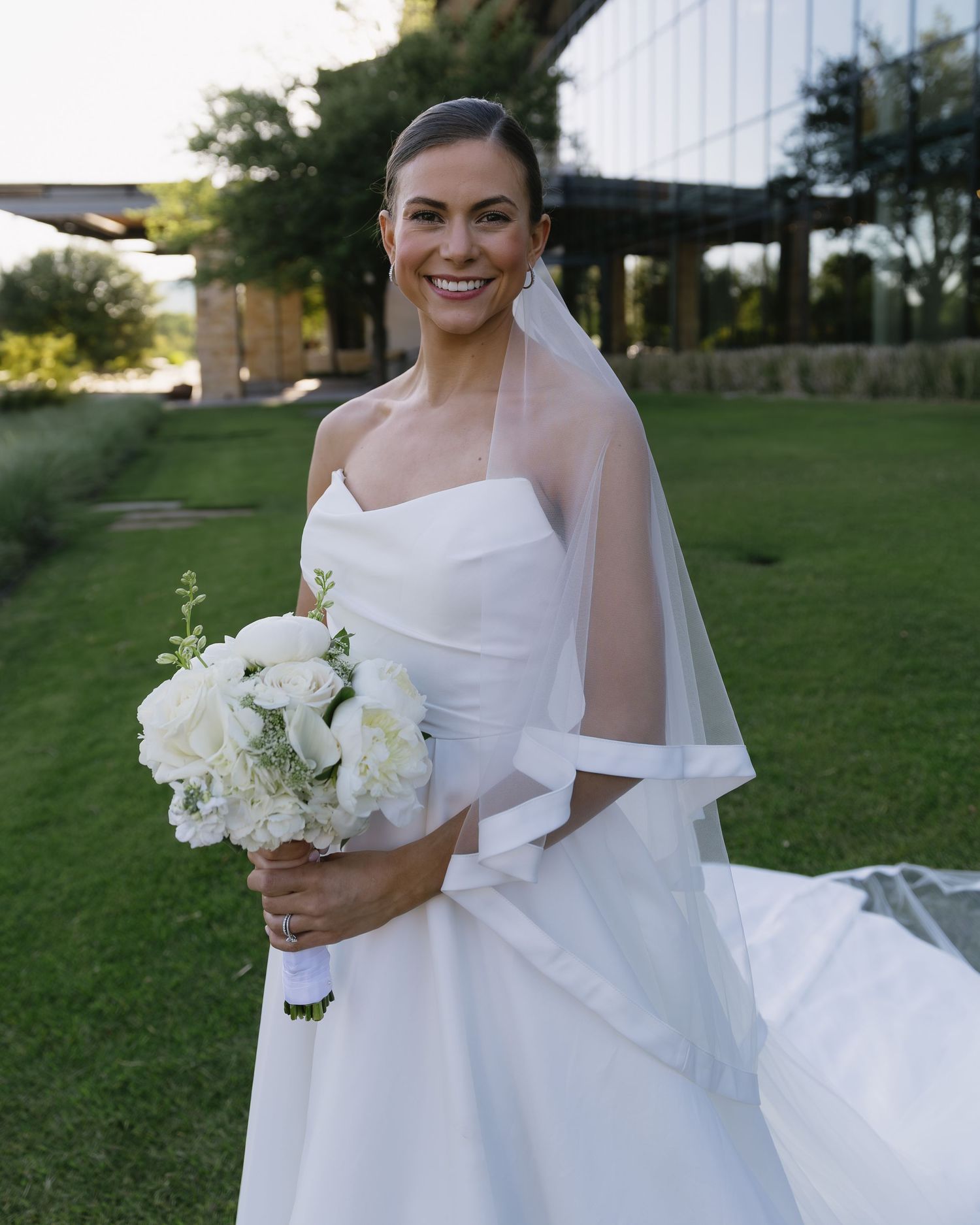 Elegant bride in a flowing white dress poses with her bouquet in a sunlit outdoor garden setting.