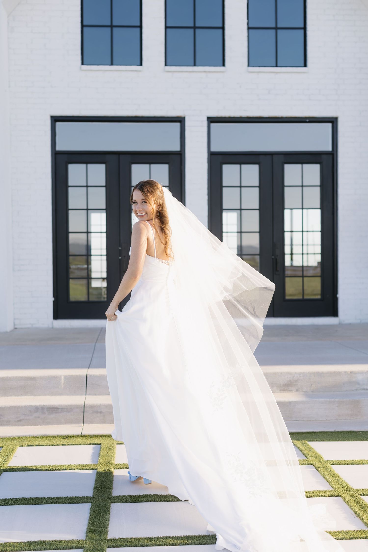 A white wedding dress and veil create dramatic movement against a modern white building with black windows.