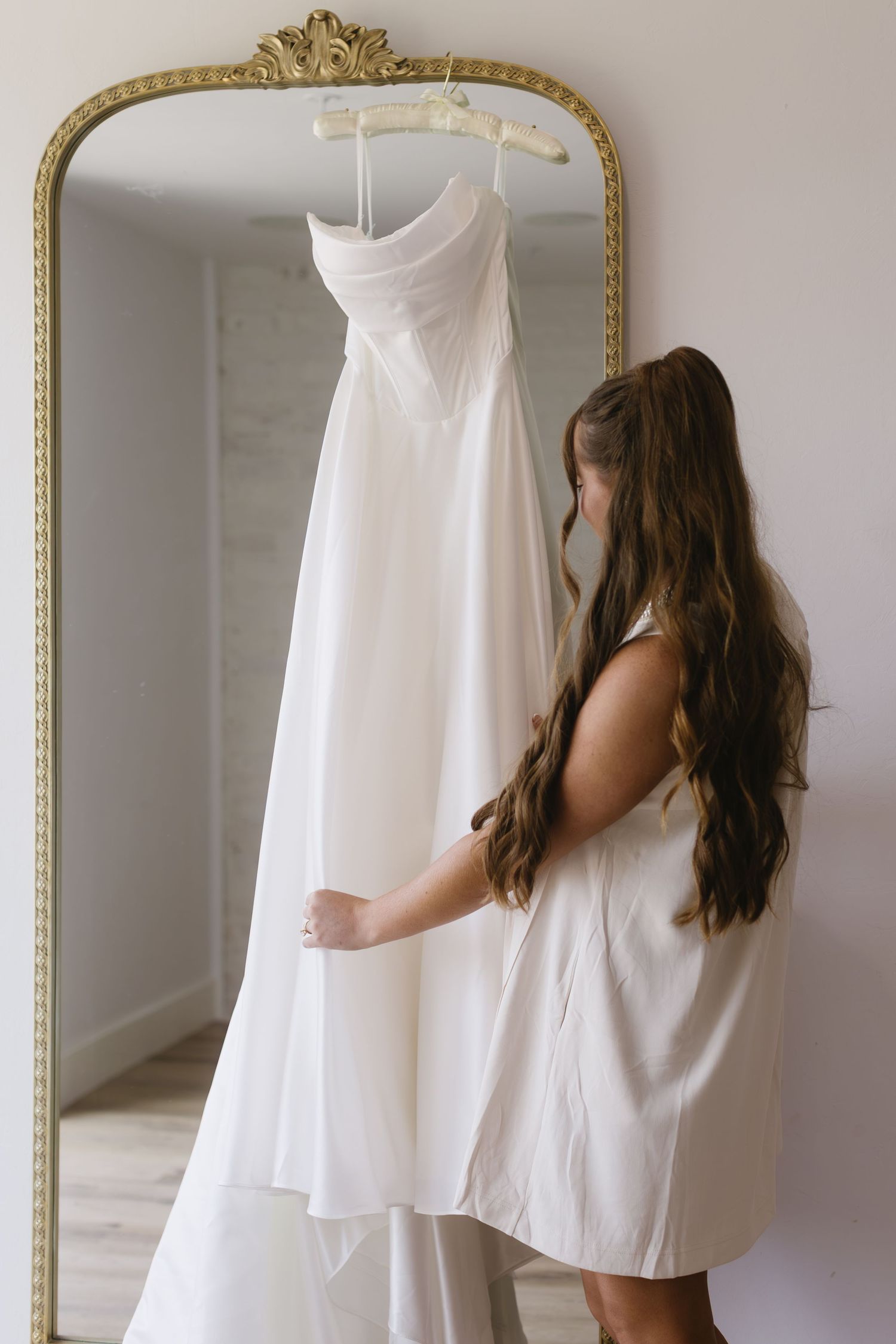 An elegant white wedding dress hangs from a vintage gold-framed mirror while someone in white admires it.