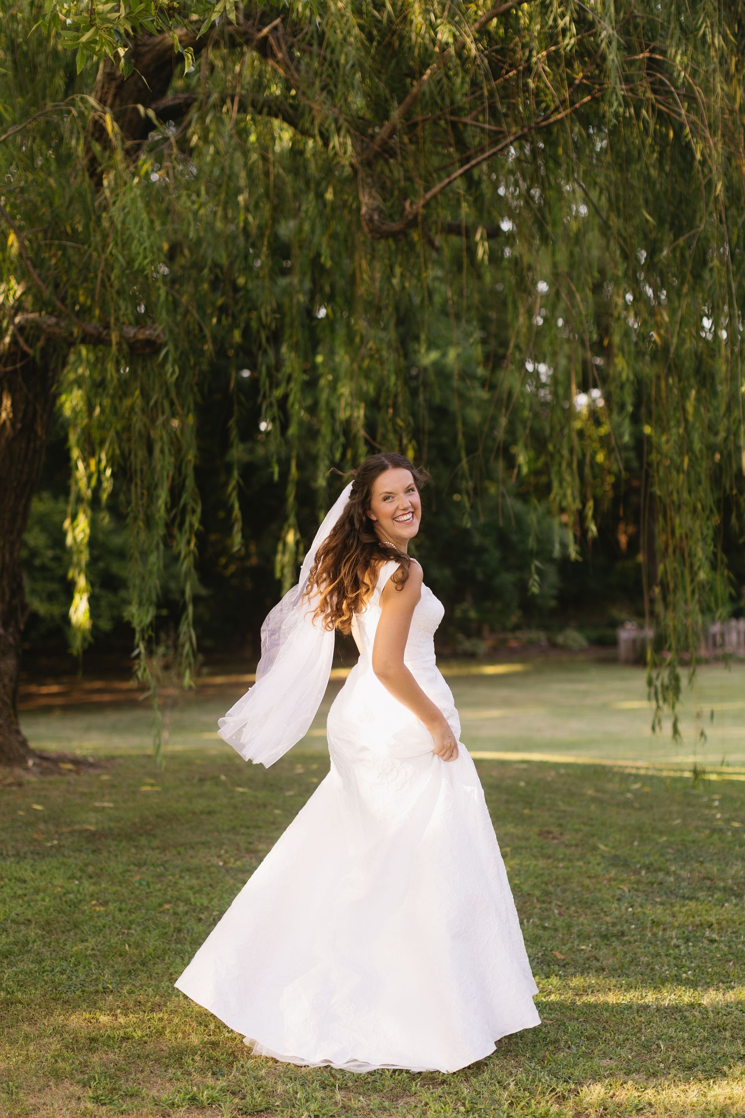 A bride in a flowing white gown twirls under a weeping willow tree.