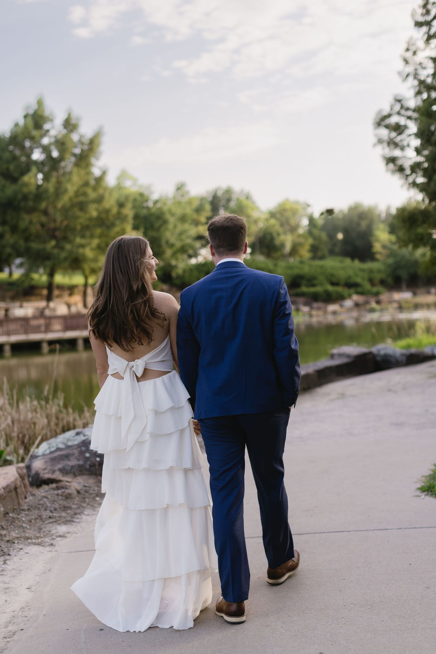 A couple in wedding attire walks along a lakeside path during golden hour.