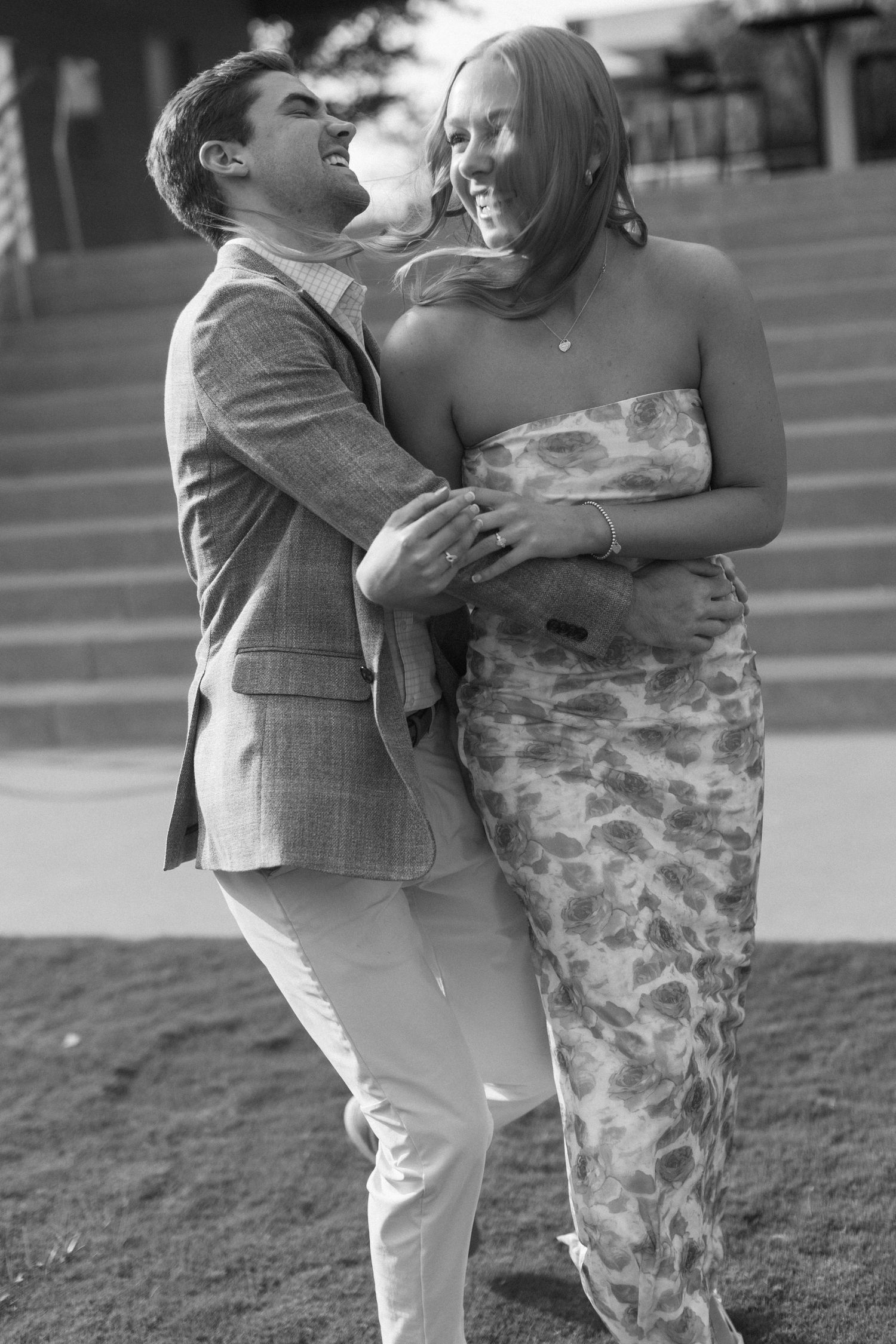 A couple in a blue floral dress and gray blazer share playful moments in a black and white outdoor photoshoot.