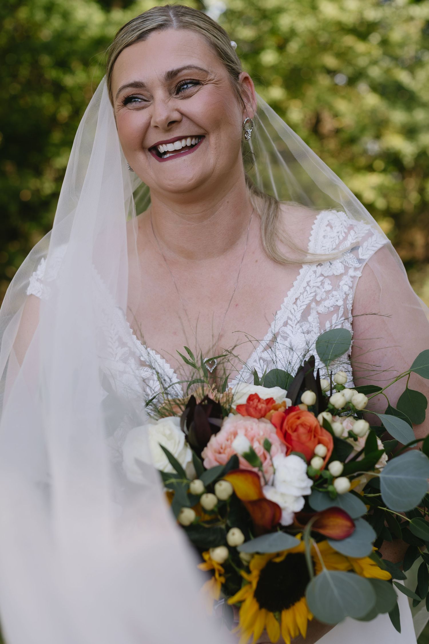 Joyful bride holding vibrant sunflower and coral rose bouquet outdoors.