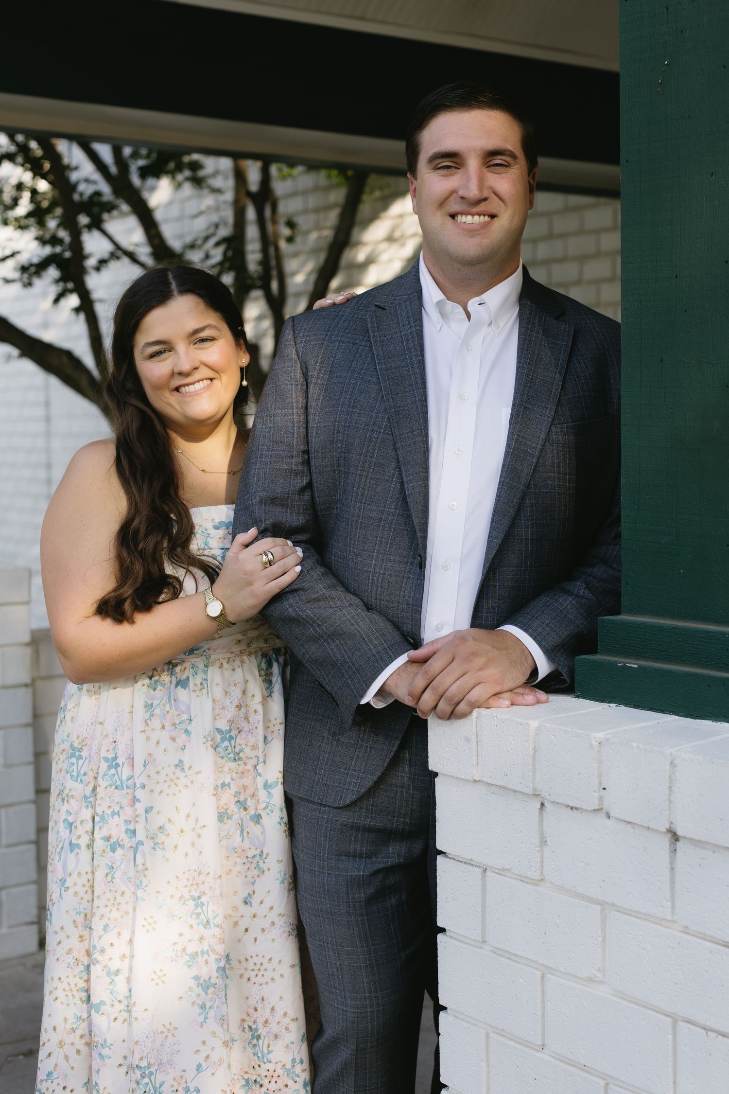 A romantic engagement photo session against a white brick wall featuring a floral dress and gray suit.
