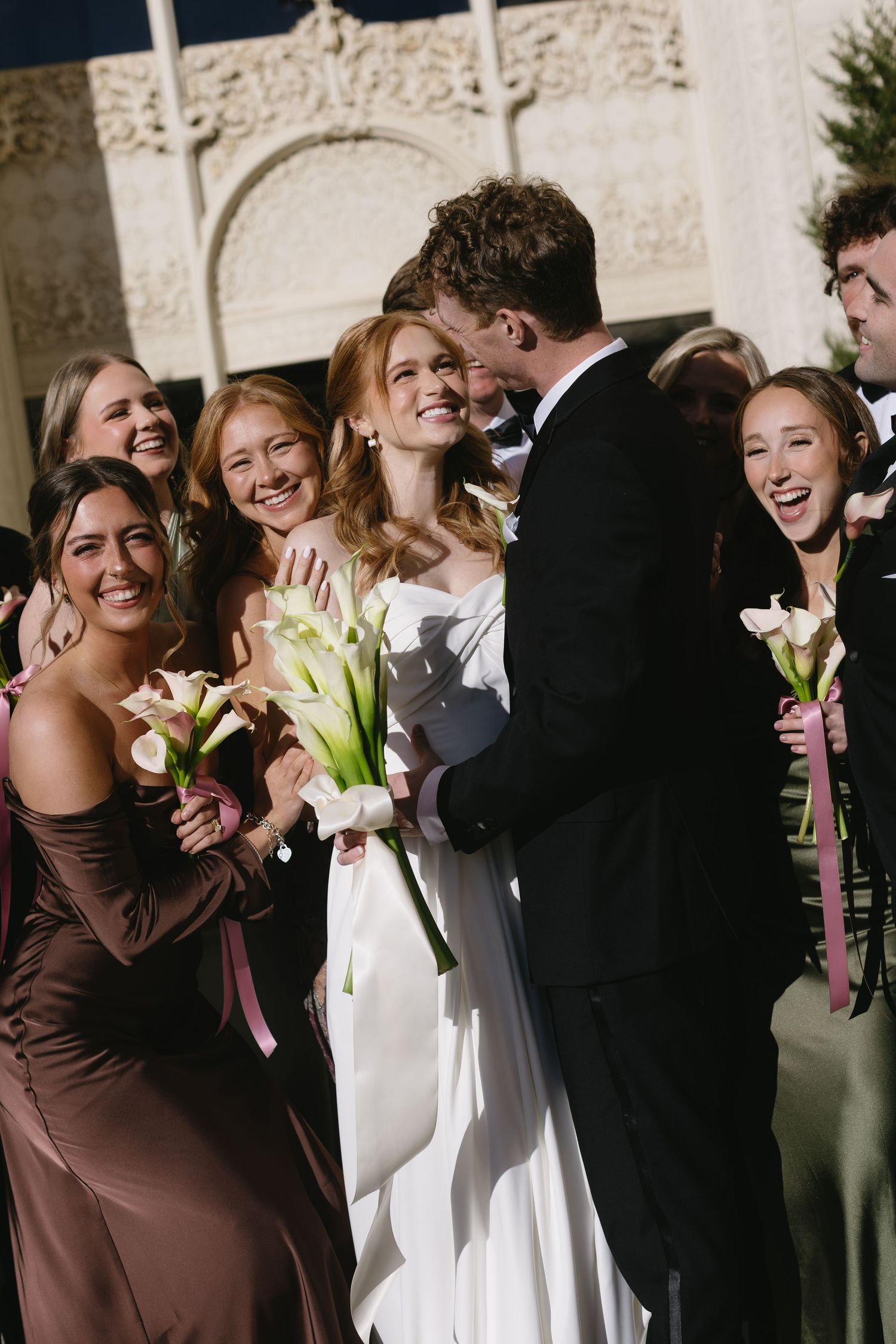 A joyful wedding party moment with bridesmaids in mixed-tone dresses holding white lilies.