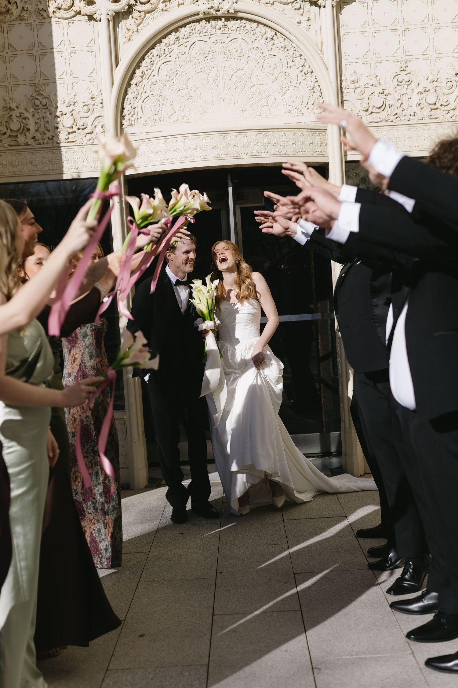 Joyful wedding celebration with guests raising hands in front of decorative white building facade.