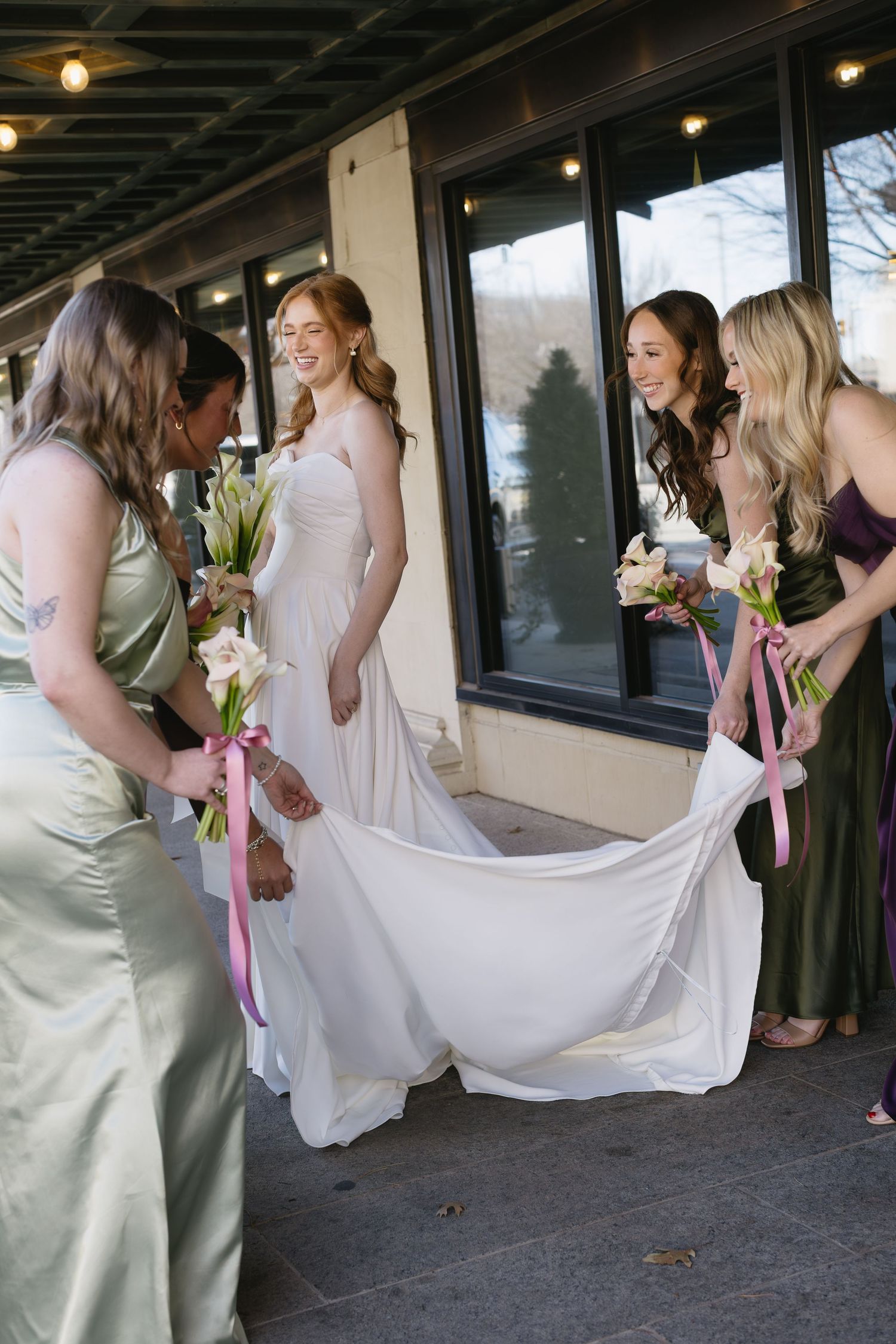Bridesmaids in various colored dresses helping with wedding dress preparations outdoors.