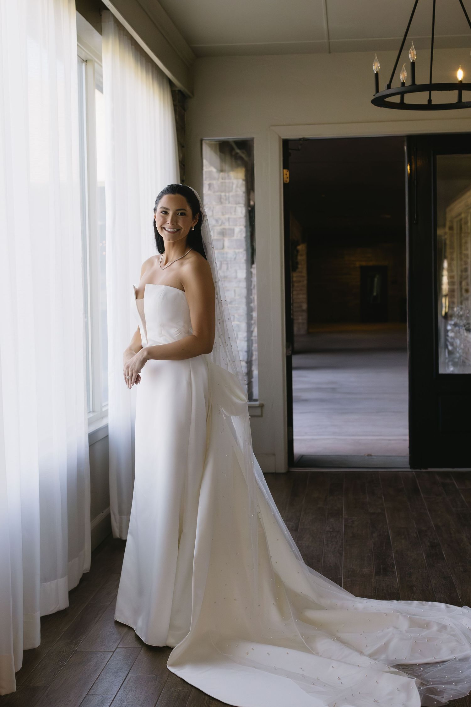 Elegant bride in white wedding dress with long train standing by window in natural light.