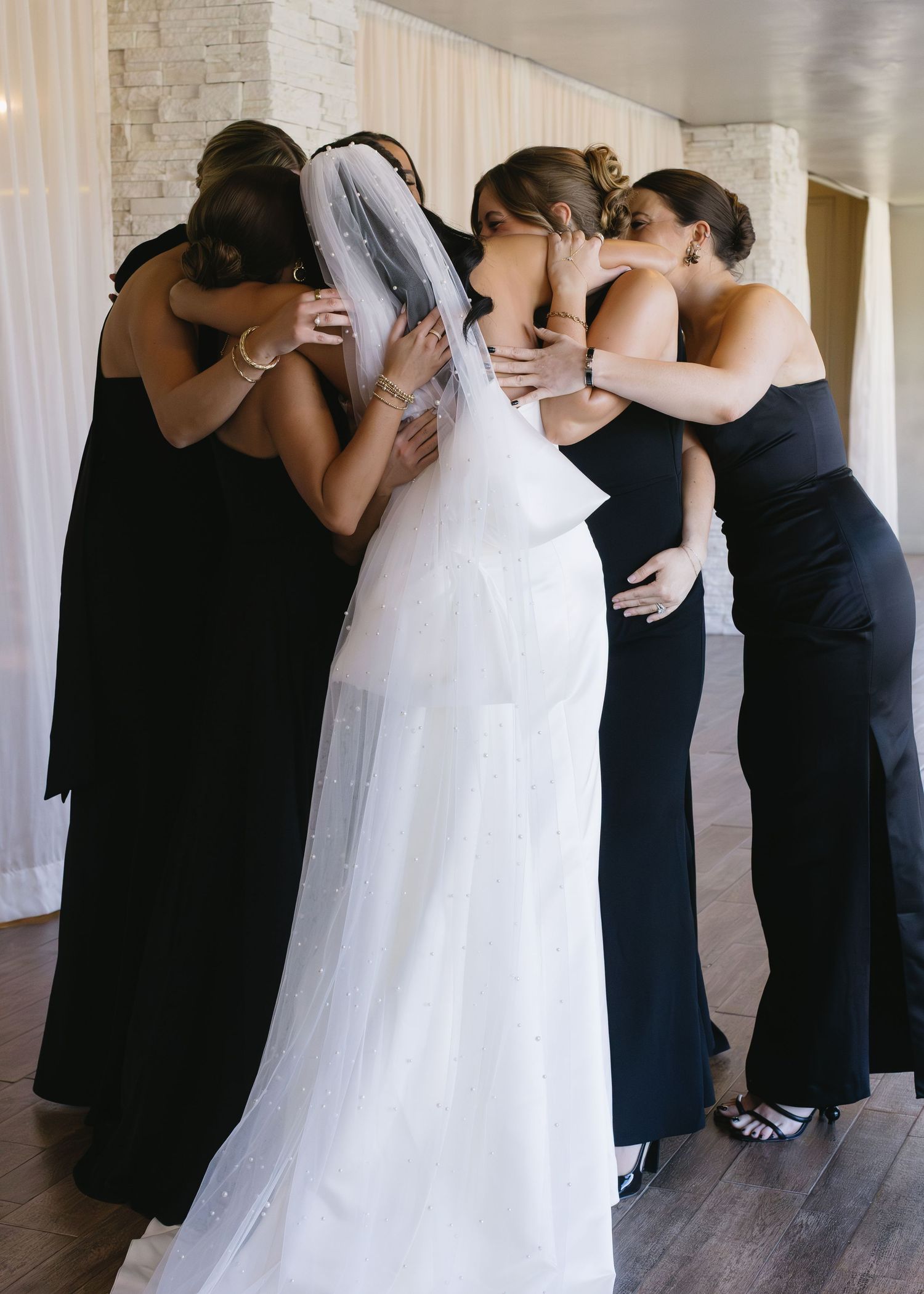 Bridesmaids in black dresses embrace bride in white gown in emotional group hug.