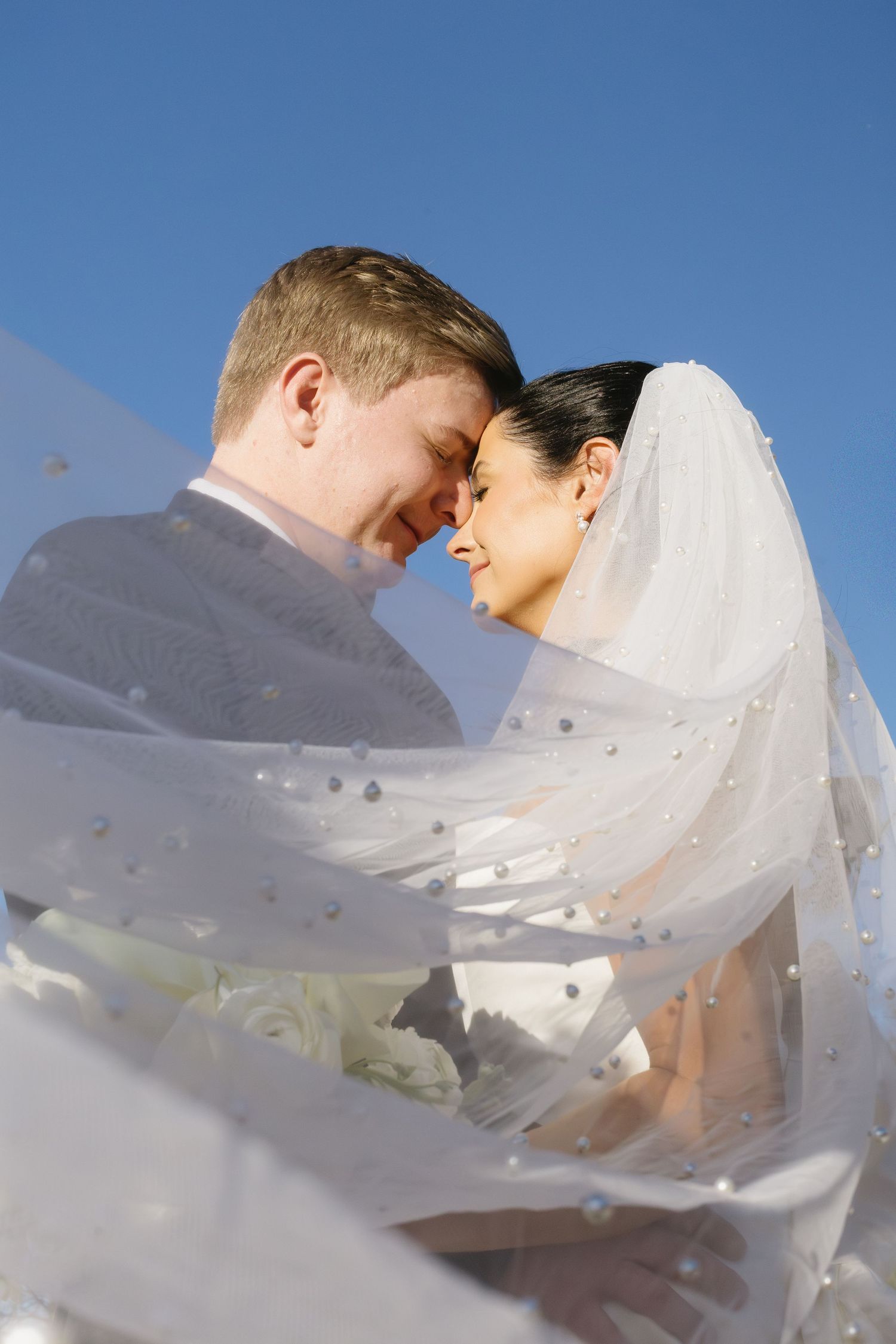 Wedding couple sharing an intimate moment under a flowing white veil against blue sky.