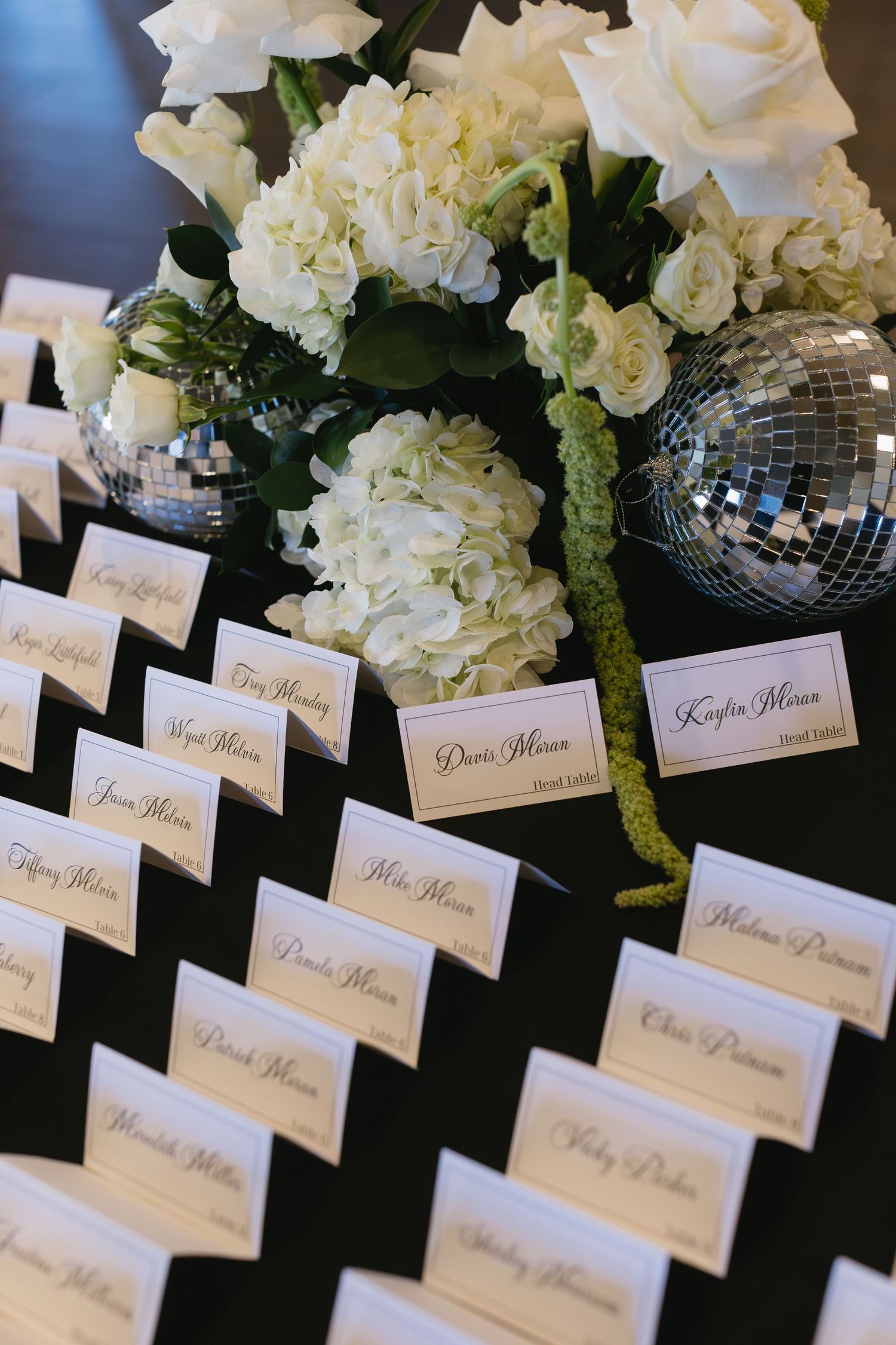 Wedding place cards arranged in rows with white floral and disco ball centerpieces.