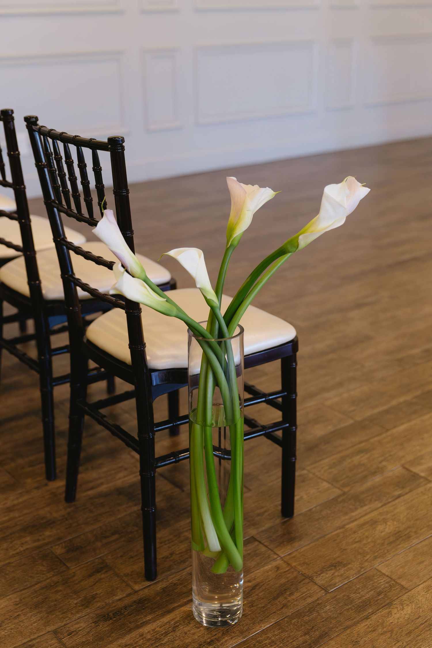 Elegant white calla lilies in glass vases arranged with black chiavari chairs.