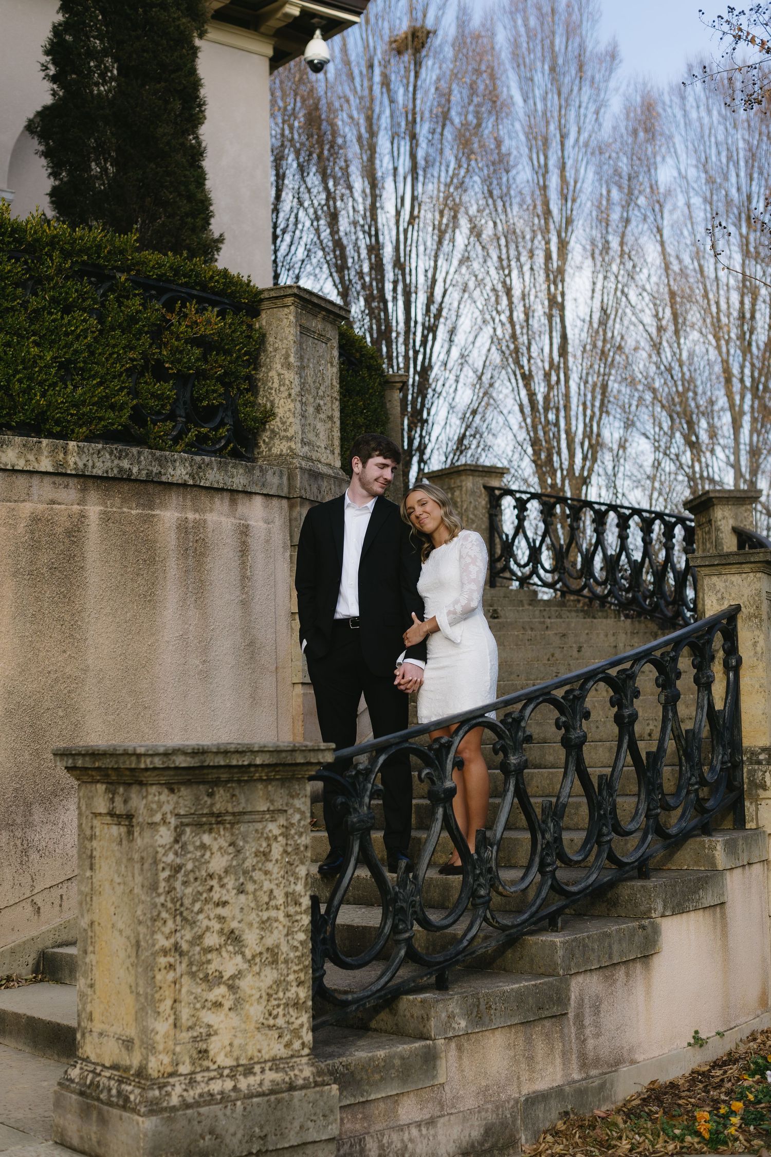 A couple stands on ornate stone steps with wrought iron railings in a romantic outdoor setting.