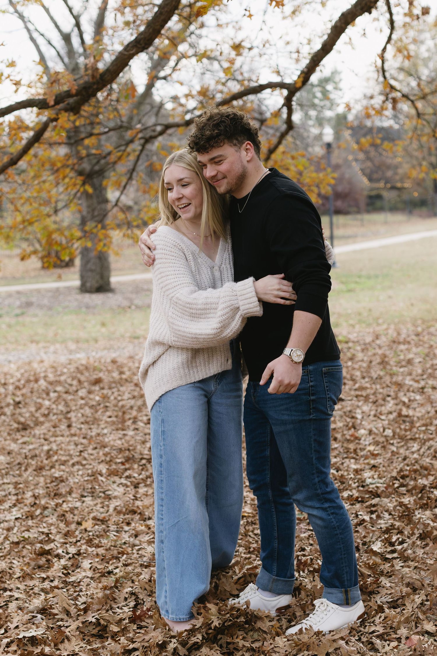 A couple embraces while standing on fallen autumn leaves under trees with golden foliage.