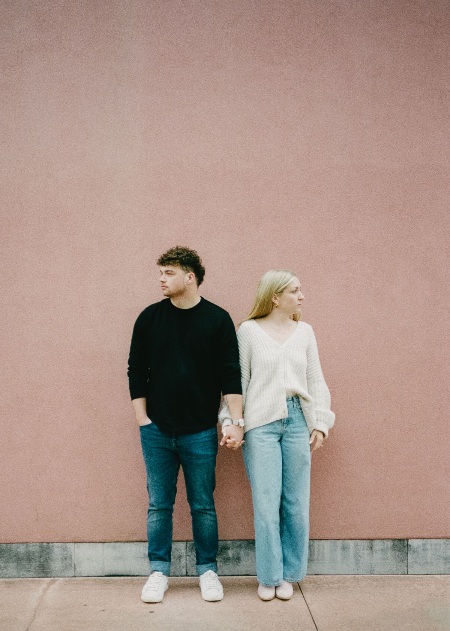 A couple holds hands while standing against a pink wall in contrasting casual outfits.