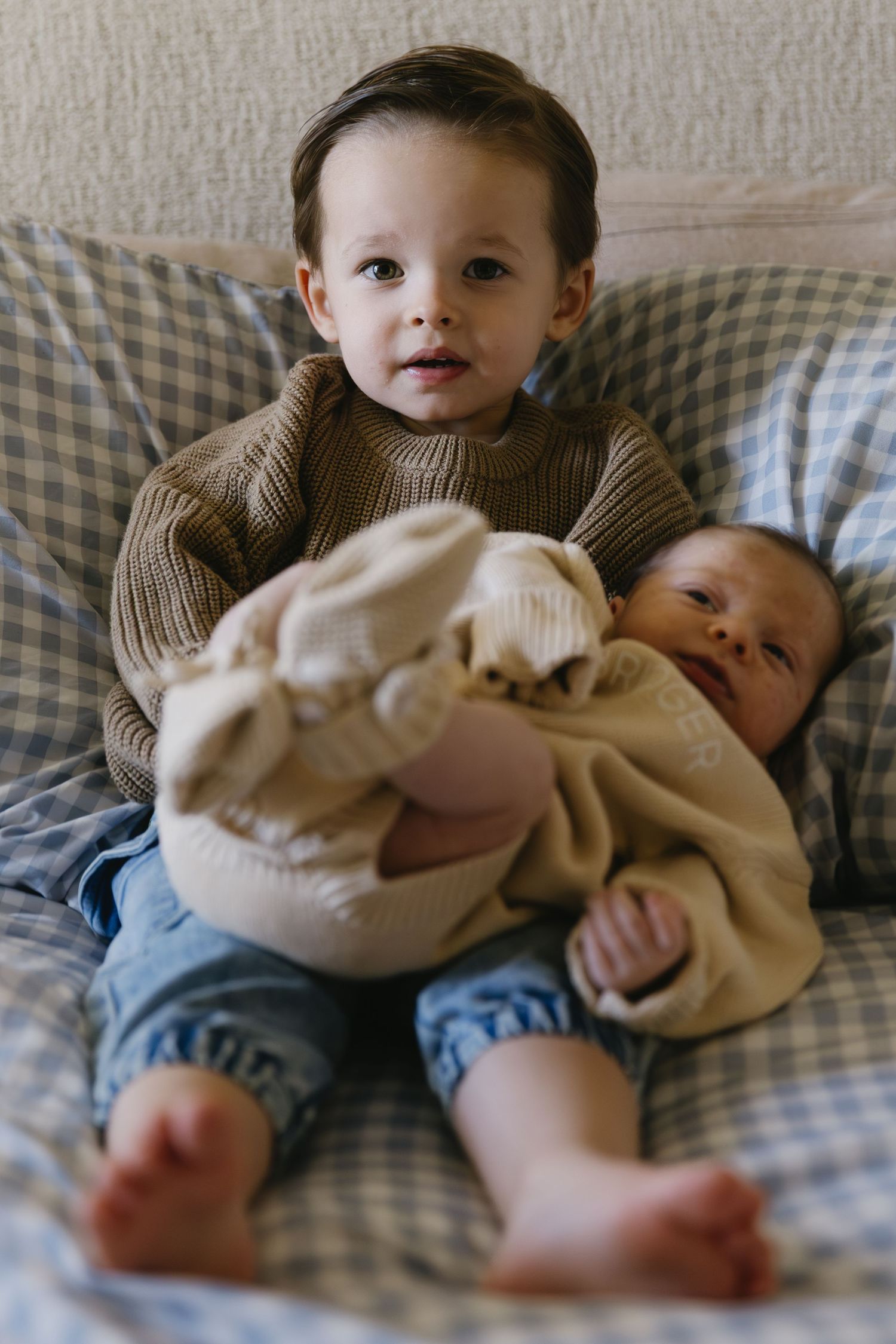 A young child in a brown sweater holds a baby on a gingham patterned couch.