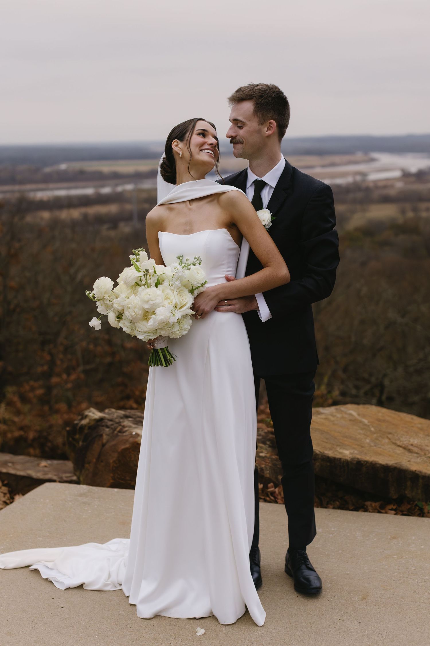 Couple in wedding attire embracing on a scenic overlook with autumn landscape.